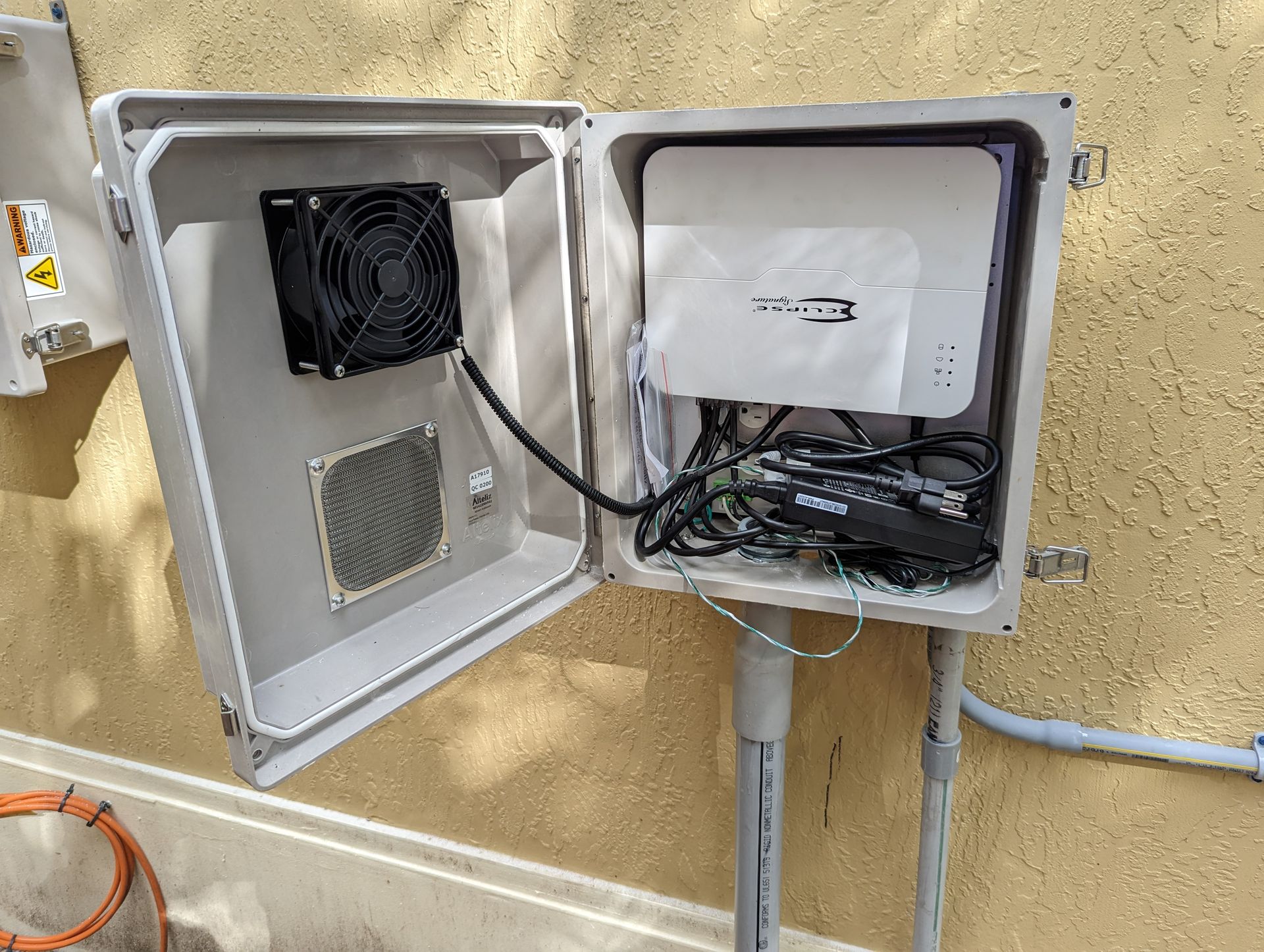Open electrical control box with router and tangled cables mounted on a yellow wall.