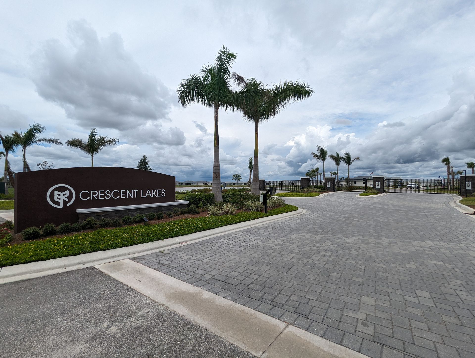 Entrance sign for Crescent Lakes with palm trees, paved roadway, and cloudy sky