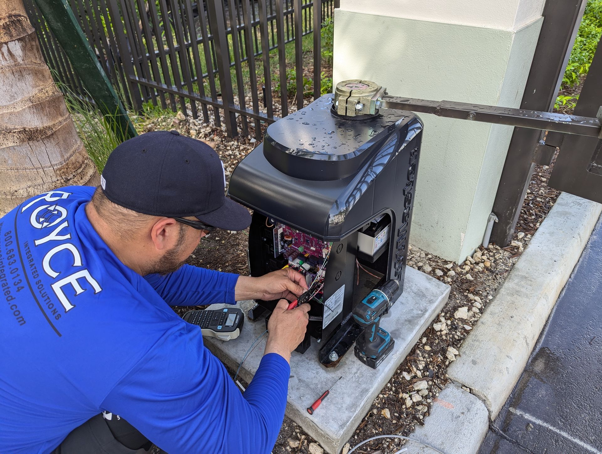 Worker in blue shirt repairs a black metal gate opener outdoors.