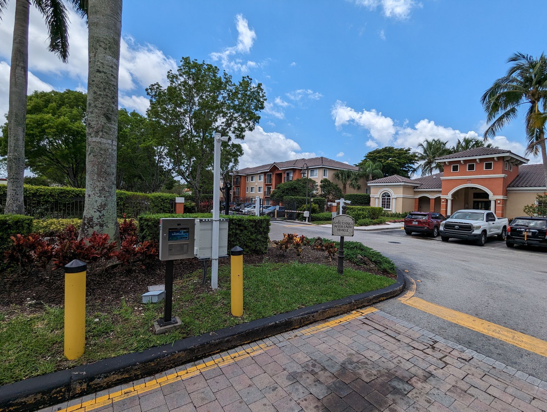 Palm-lined driveway entrance to a residential complex with parked cars and landscaped flower beds under a blue sky