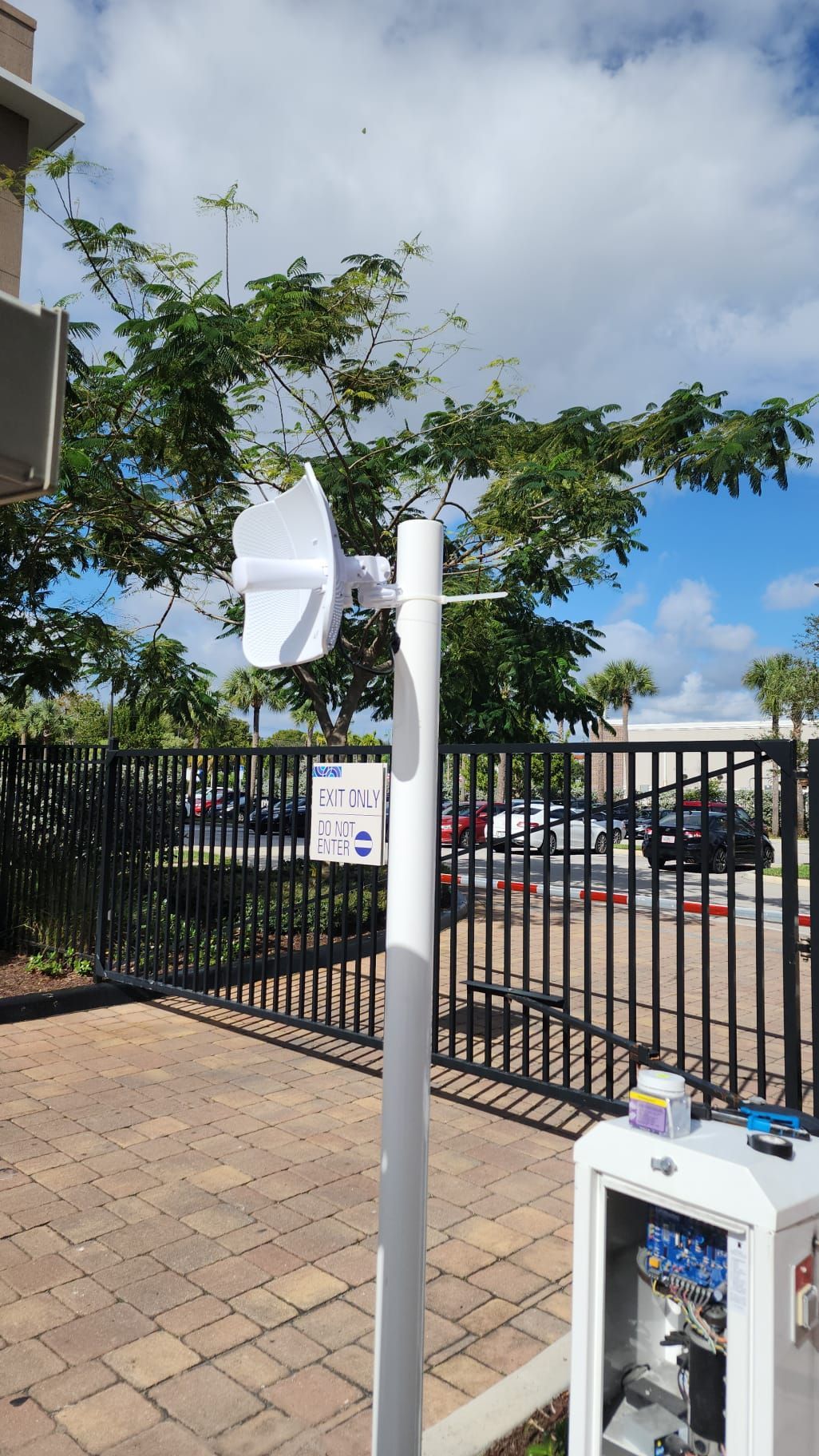 White weather sensor on a pole beside a parking lot and black fence under a blue sky.