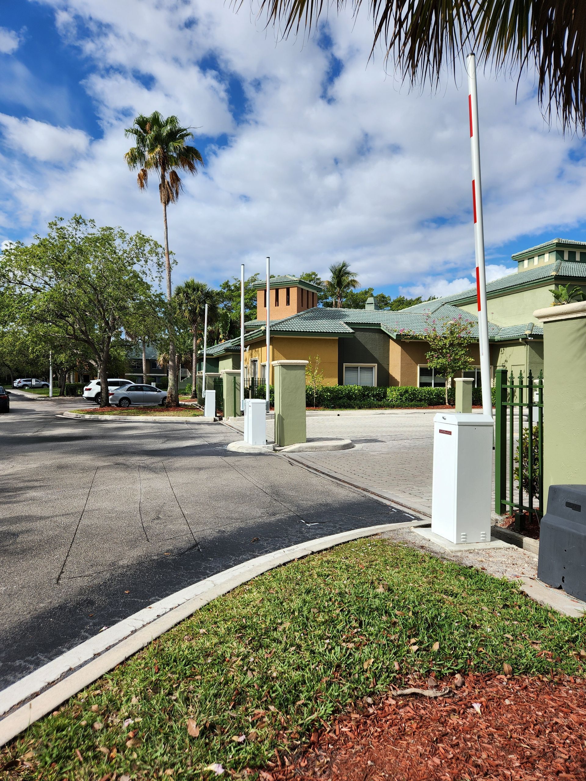 Gated entrance with barrier arm, white pillars, palm trees, and townhouses under a partly cloudy sky