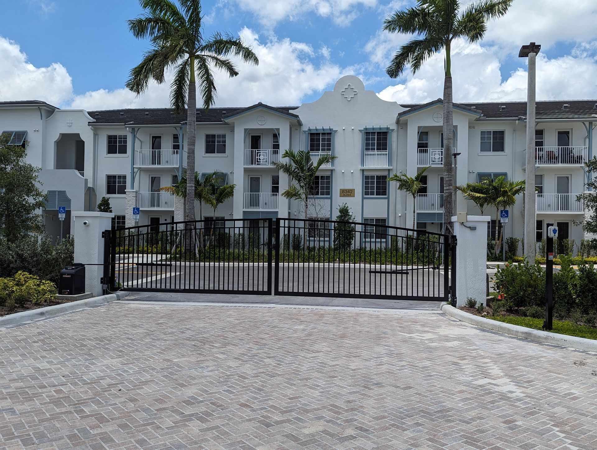 Gated white apartment building with palm trees and a cobblestone driveway under a cloudy sky