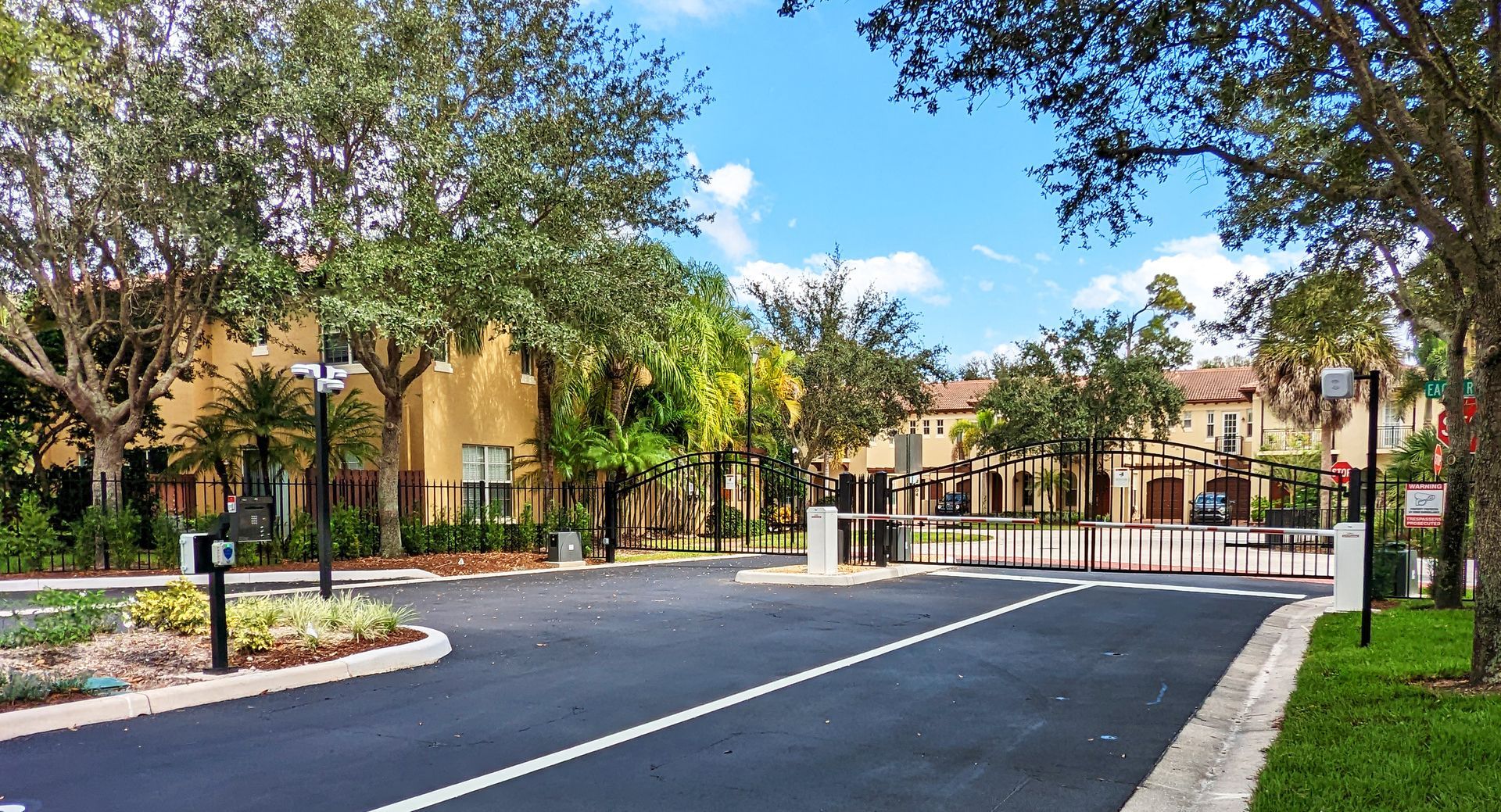 Gated residential entrance with palm trees, brick columns, and a paved driveway under a blue sky