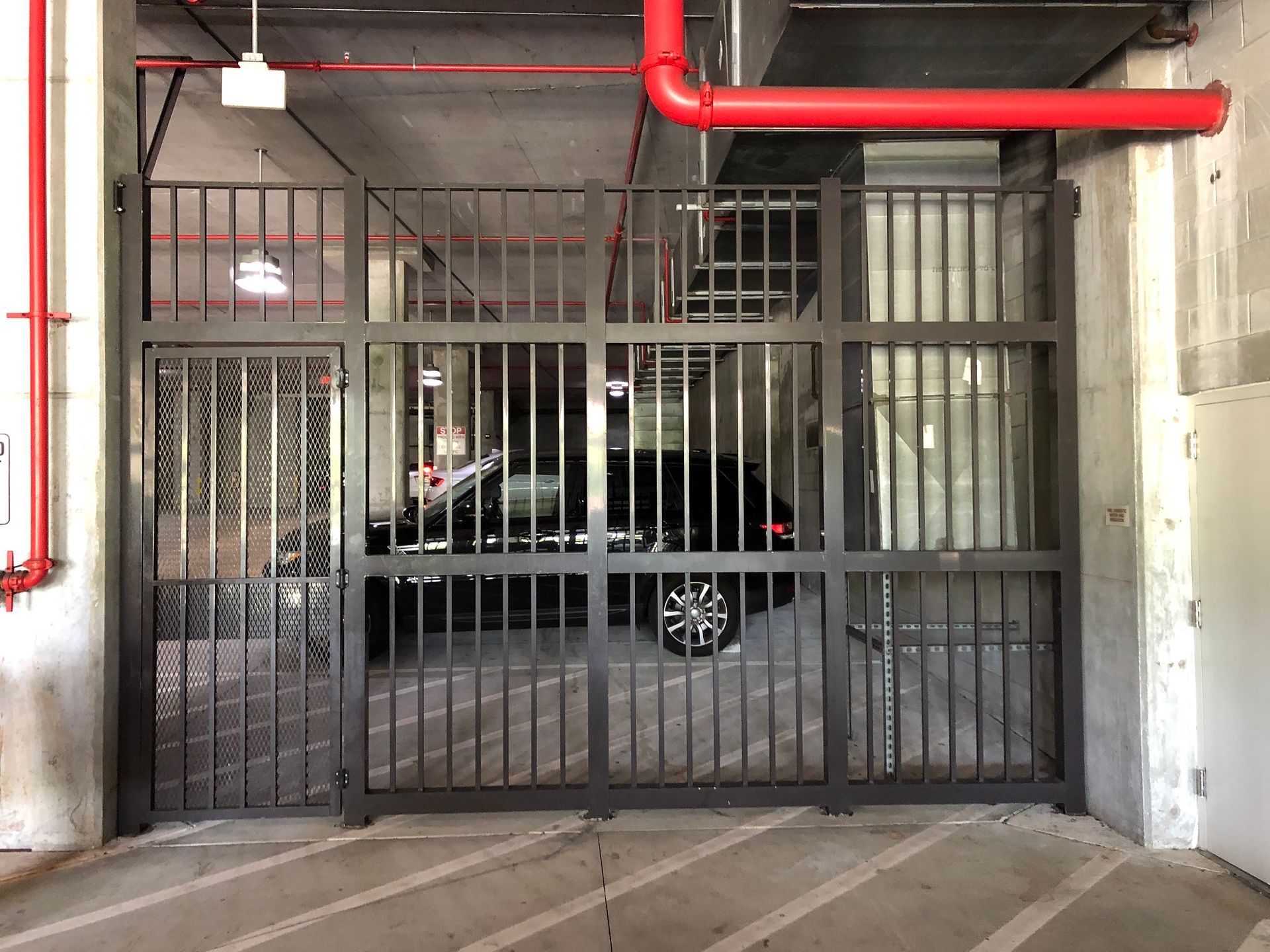 Black car behind a closed metal gate in a concrete parking garage with red pipes overhead