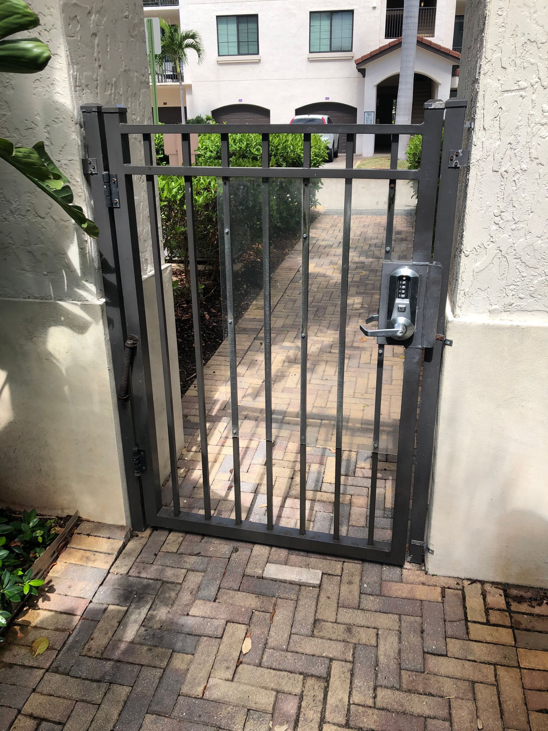 Black metal gate with lock at a courtyard entrance, bordered by stucco walls and brick pavers.