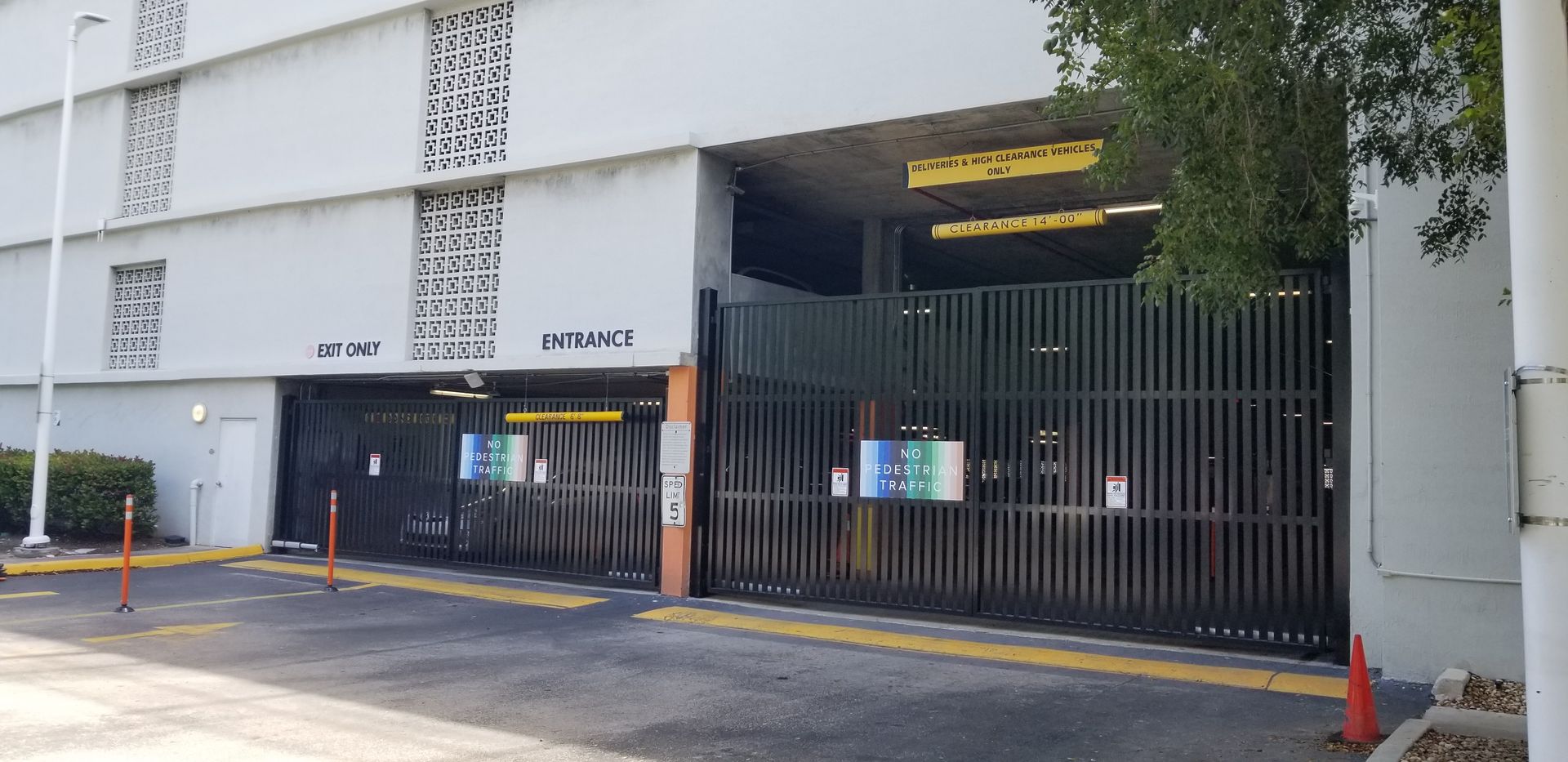 Gated parking garage entrance beside a white building, with orange cones and a small entrance booth.