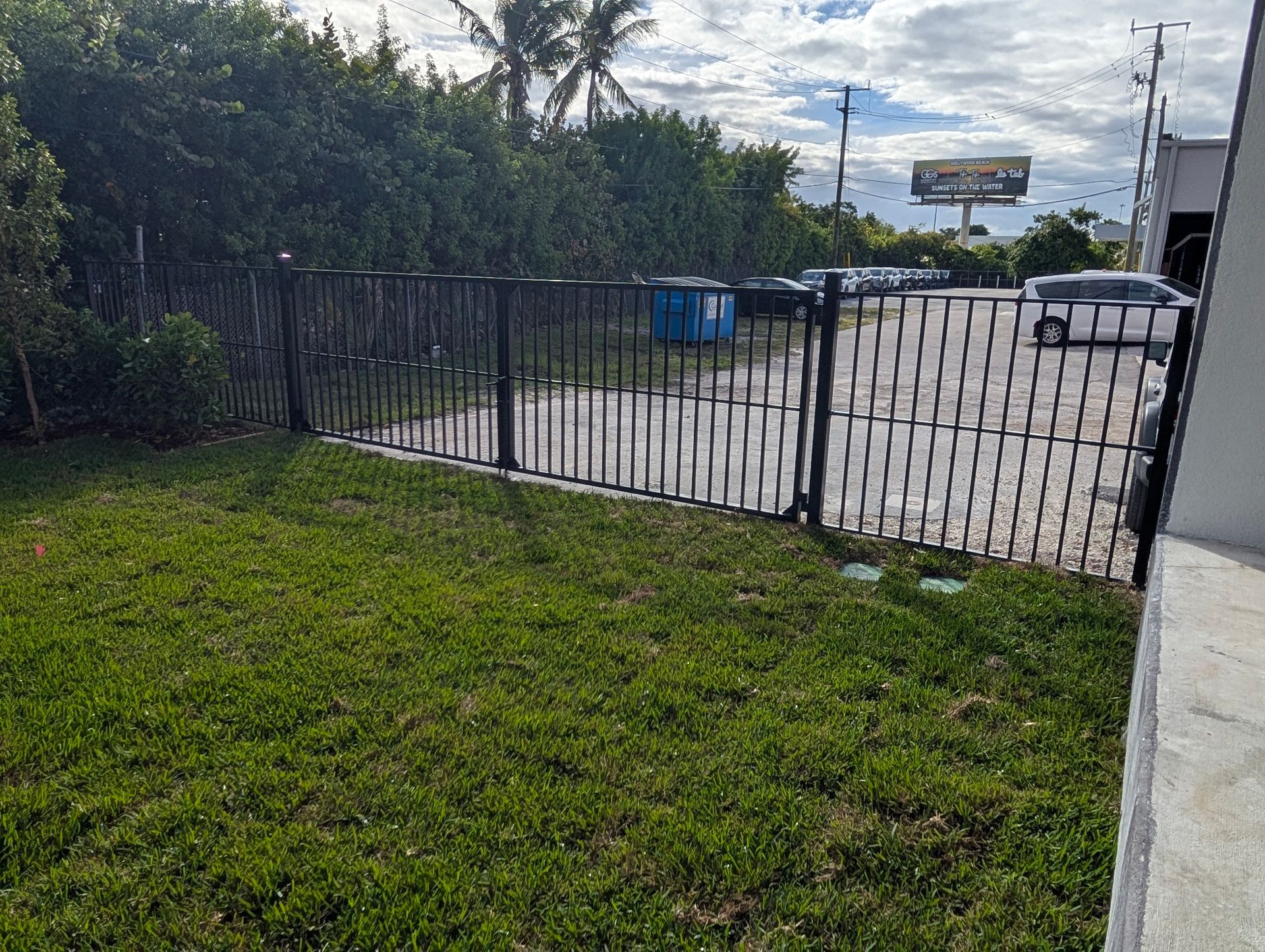 Grassy yard beside a black metal gate opening to a paved lot with trees and a parked car.