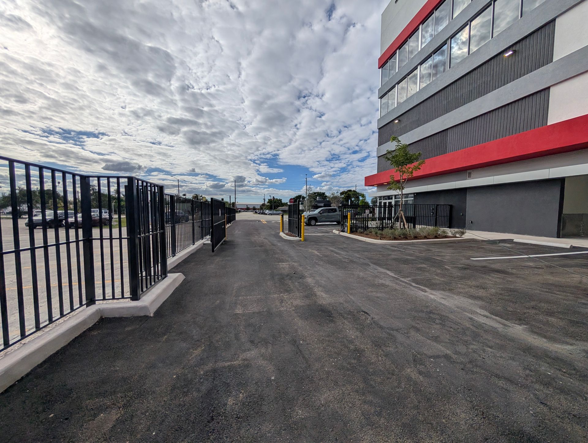 Empty parking lot beside a black fence and a gray building with red stripes under a cloudy sky