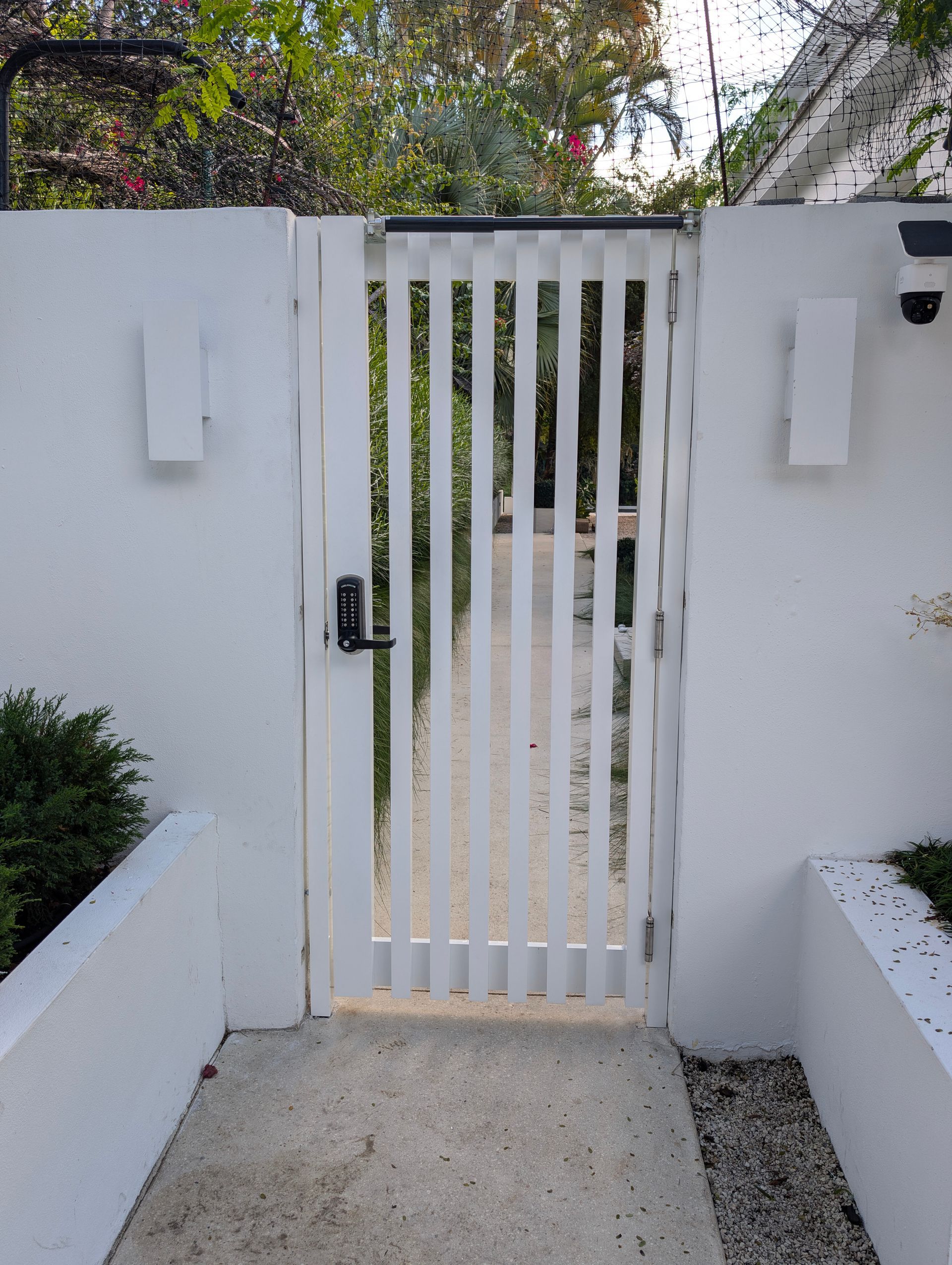 White slatted pedestrian gate in a concrete wall with a keypad lock, opening to a garden path.