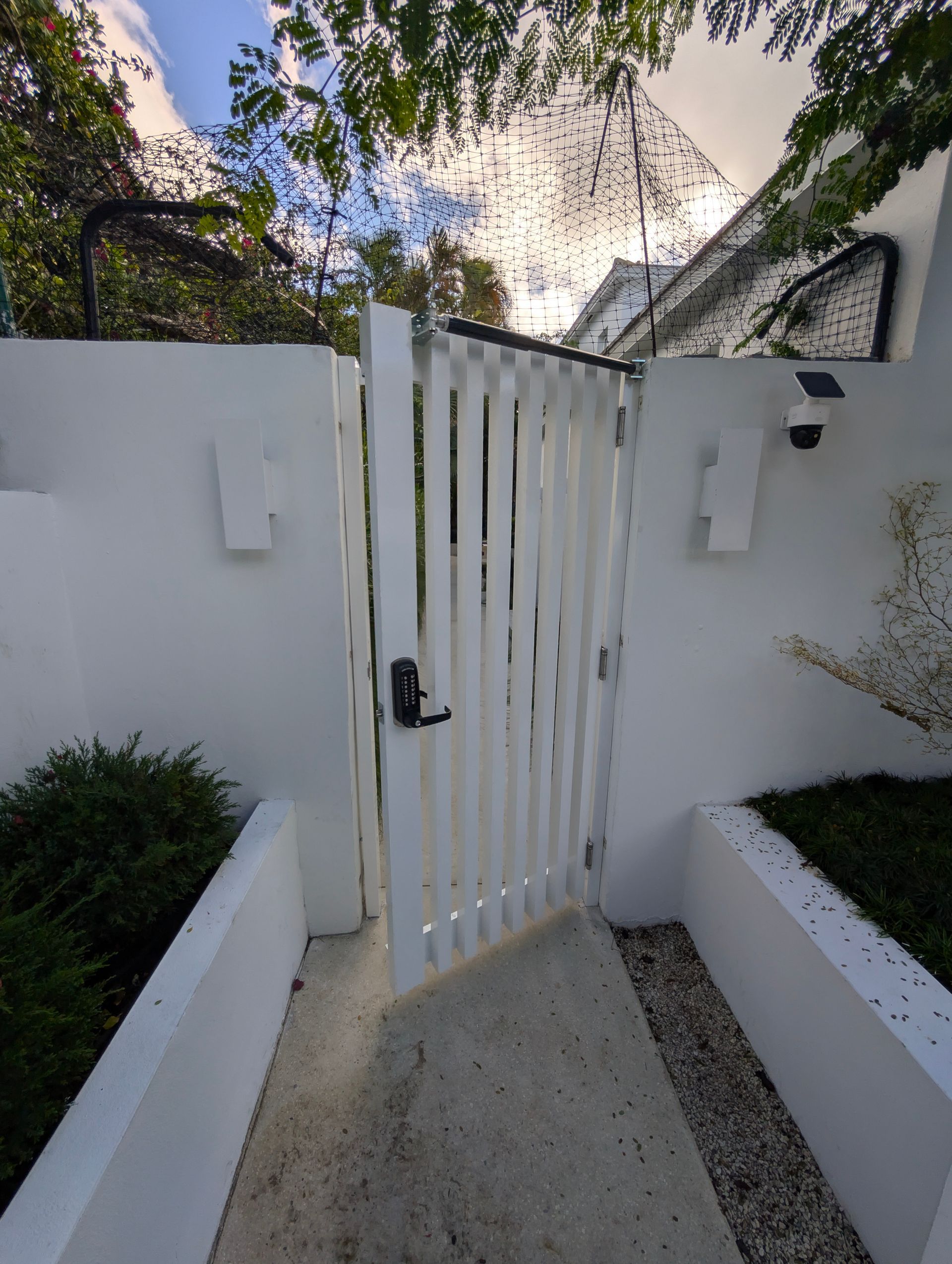 White front gate between stucco walls, with narrow concrete path and greenery on both sides.
