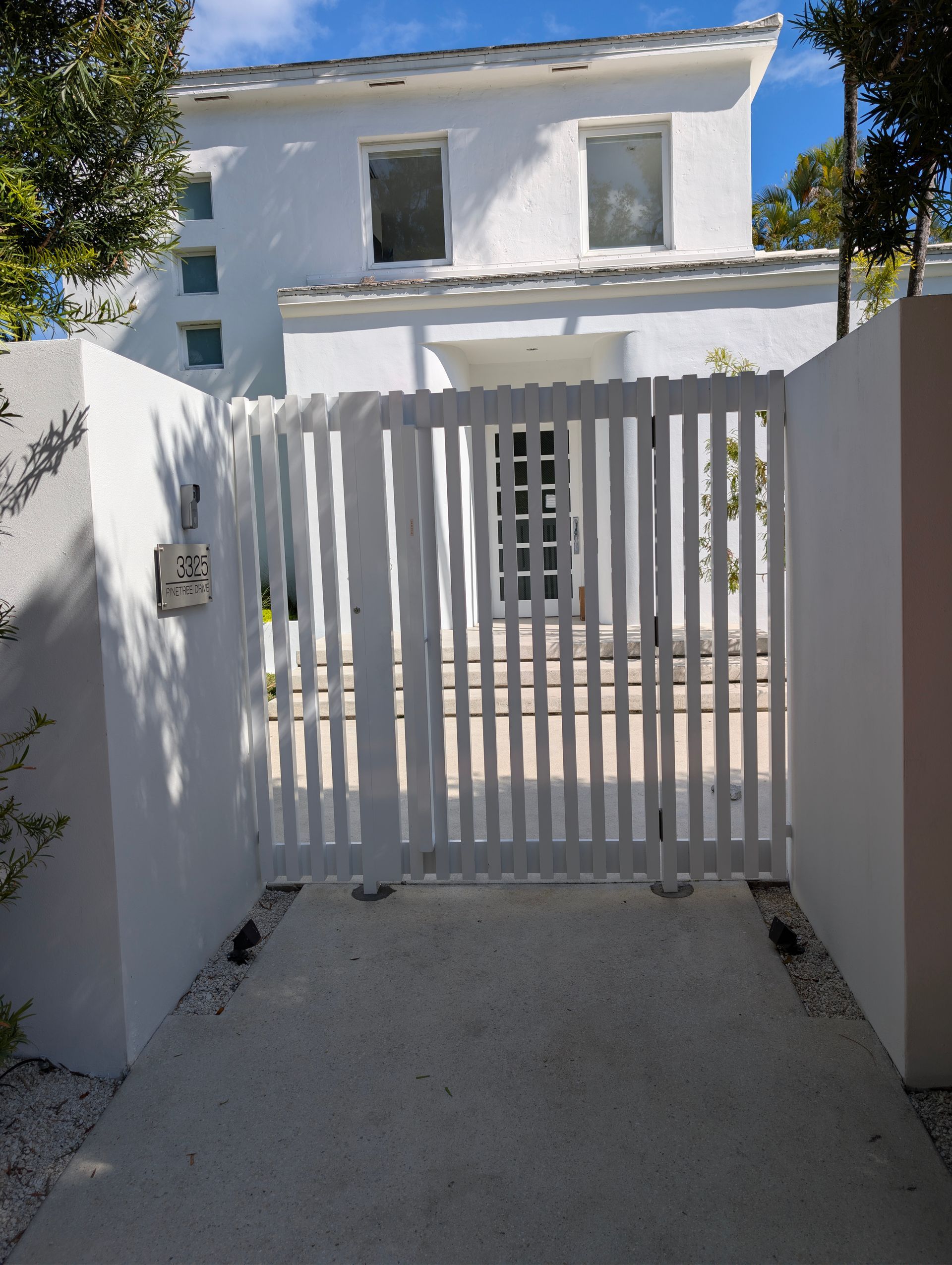White house seen through a closed white slatted gate at the end of a narrow concrete walkway.