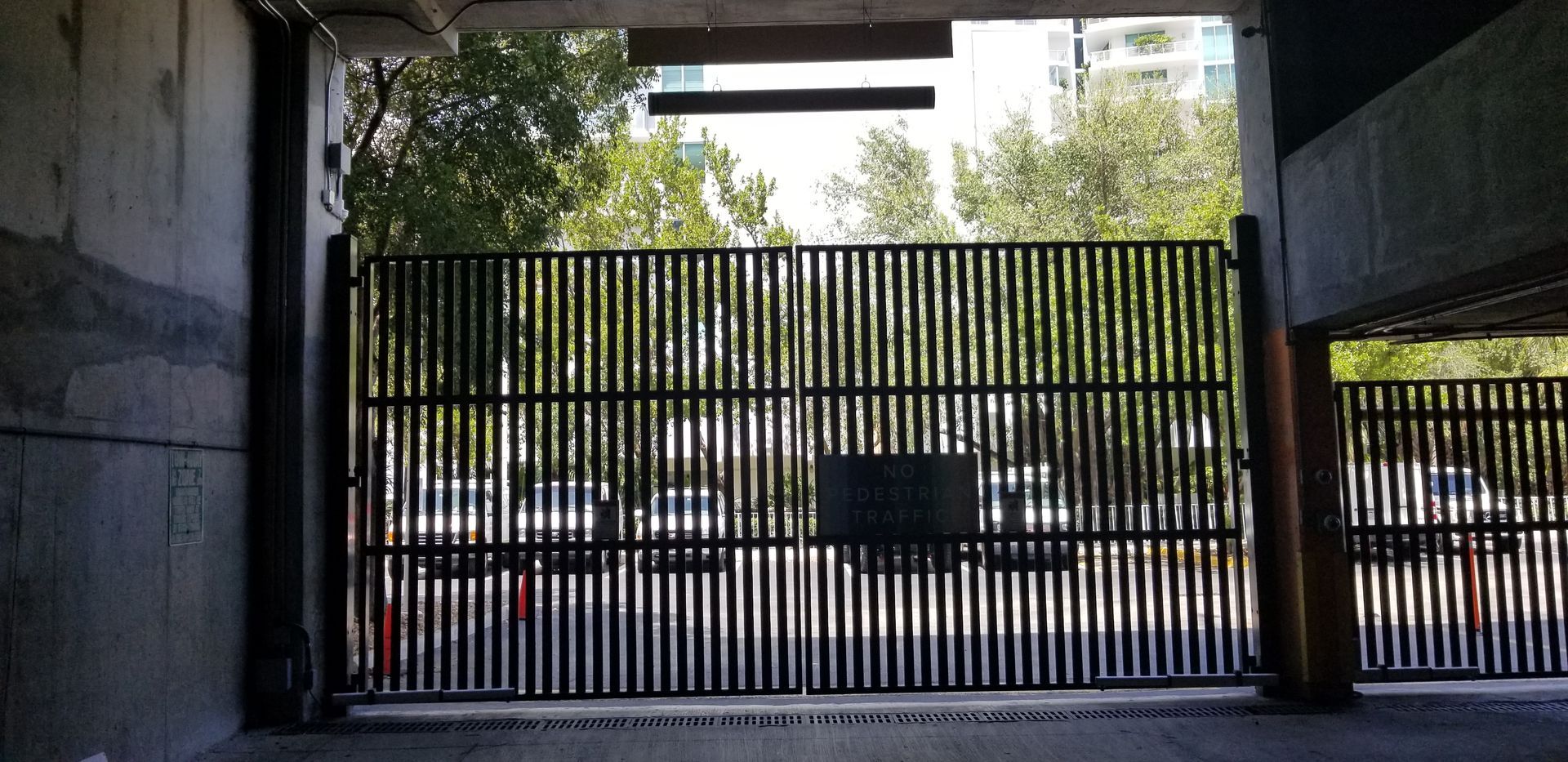 Shaded parking area with a black metal gate, parked cars, and trees beyond the entrance.