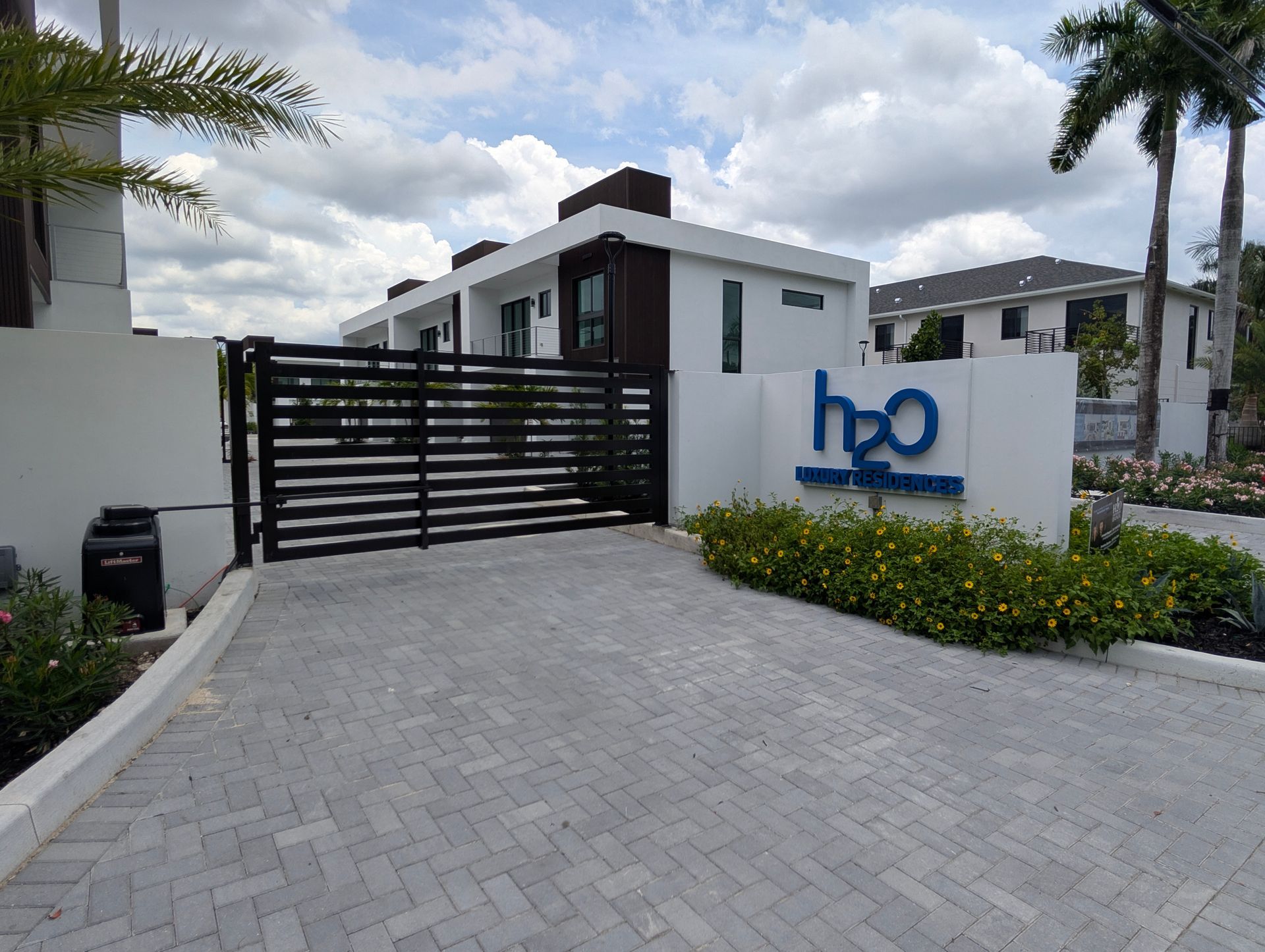 Modern gated entrance with black gate, white walls, blue logo sign, and a paved driveway