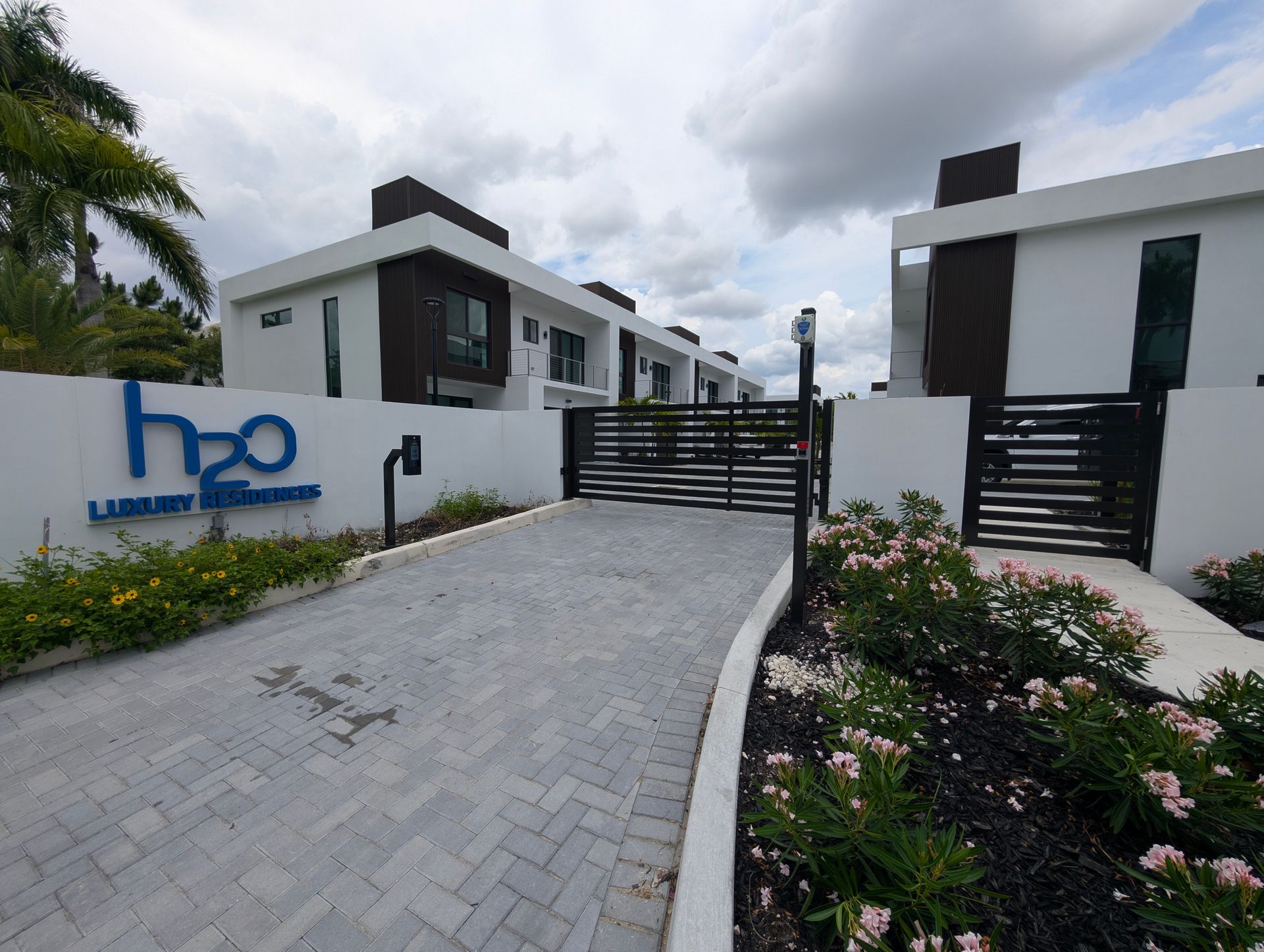 Modern white apartment complex entrance with black gates, driveway, and flower beds under a cloudy sky