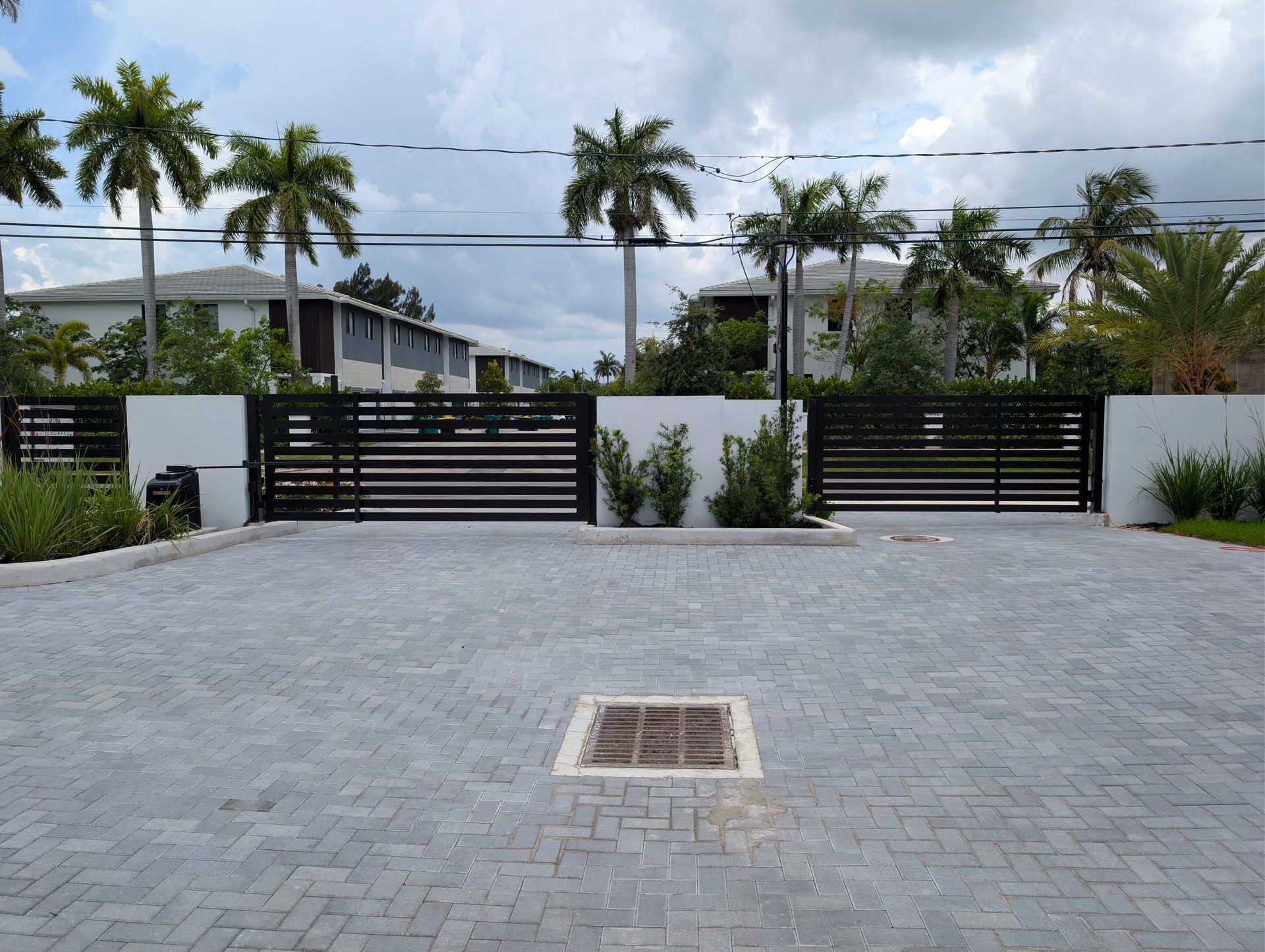 Modern house entrance with black gates, white walls, palm trees, and a paved driveway under cloudy skies