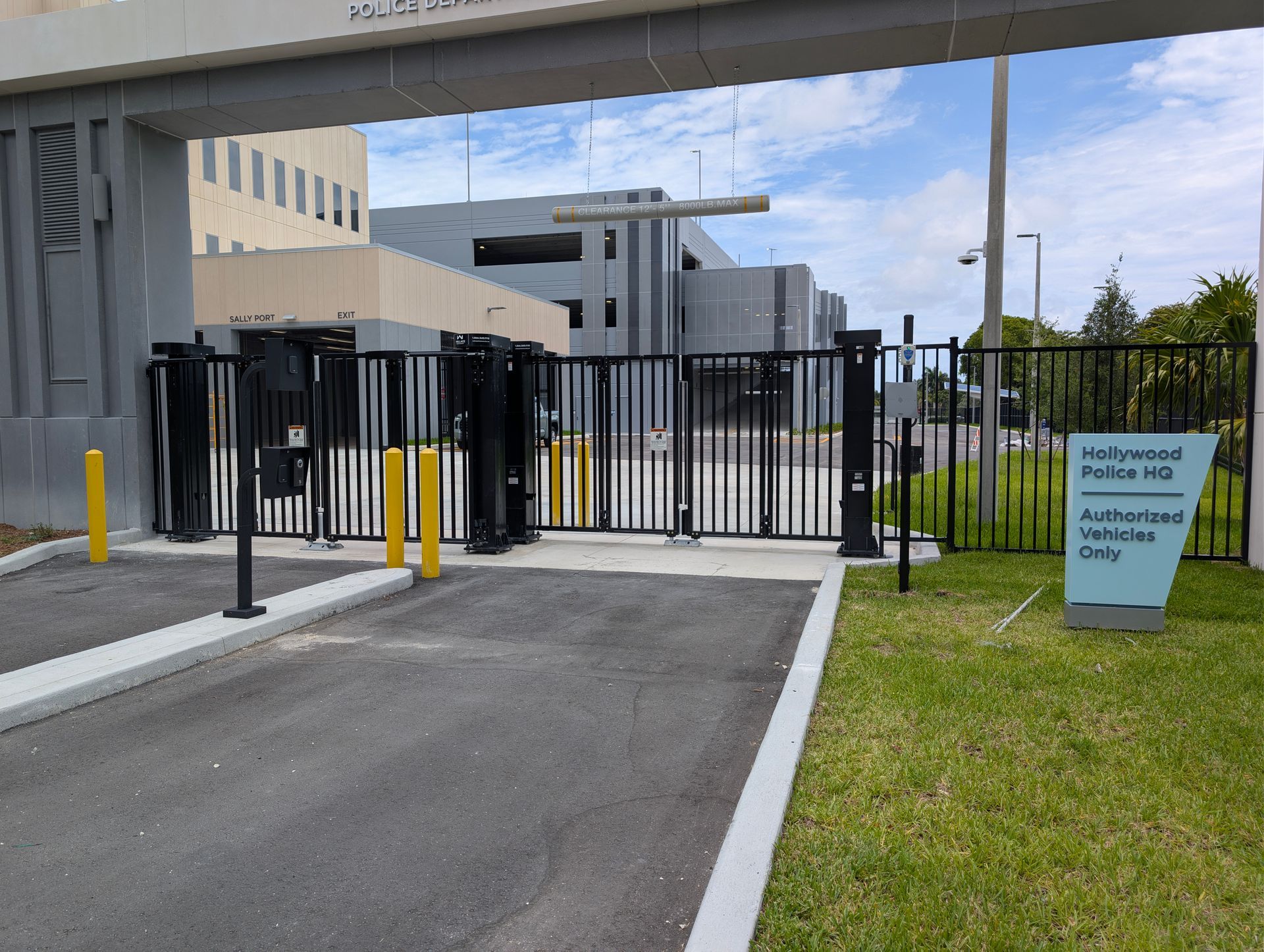 Gated entrance with black fence and bollards beside a driveway and grassy area under an overcast sky