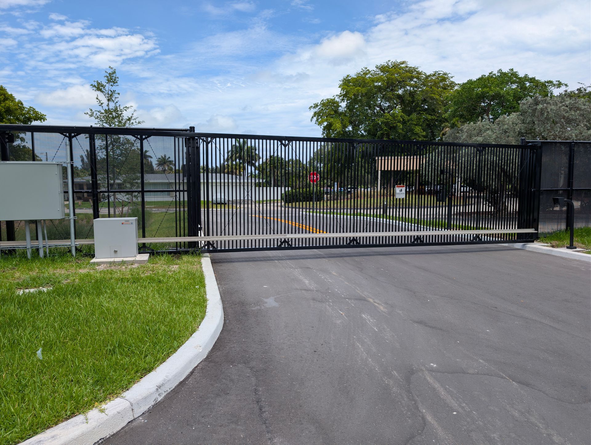 Black sliding gate across a paved driveway at a gated entrance with grass and trees beyond