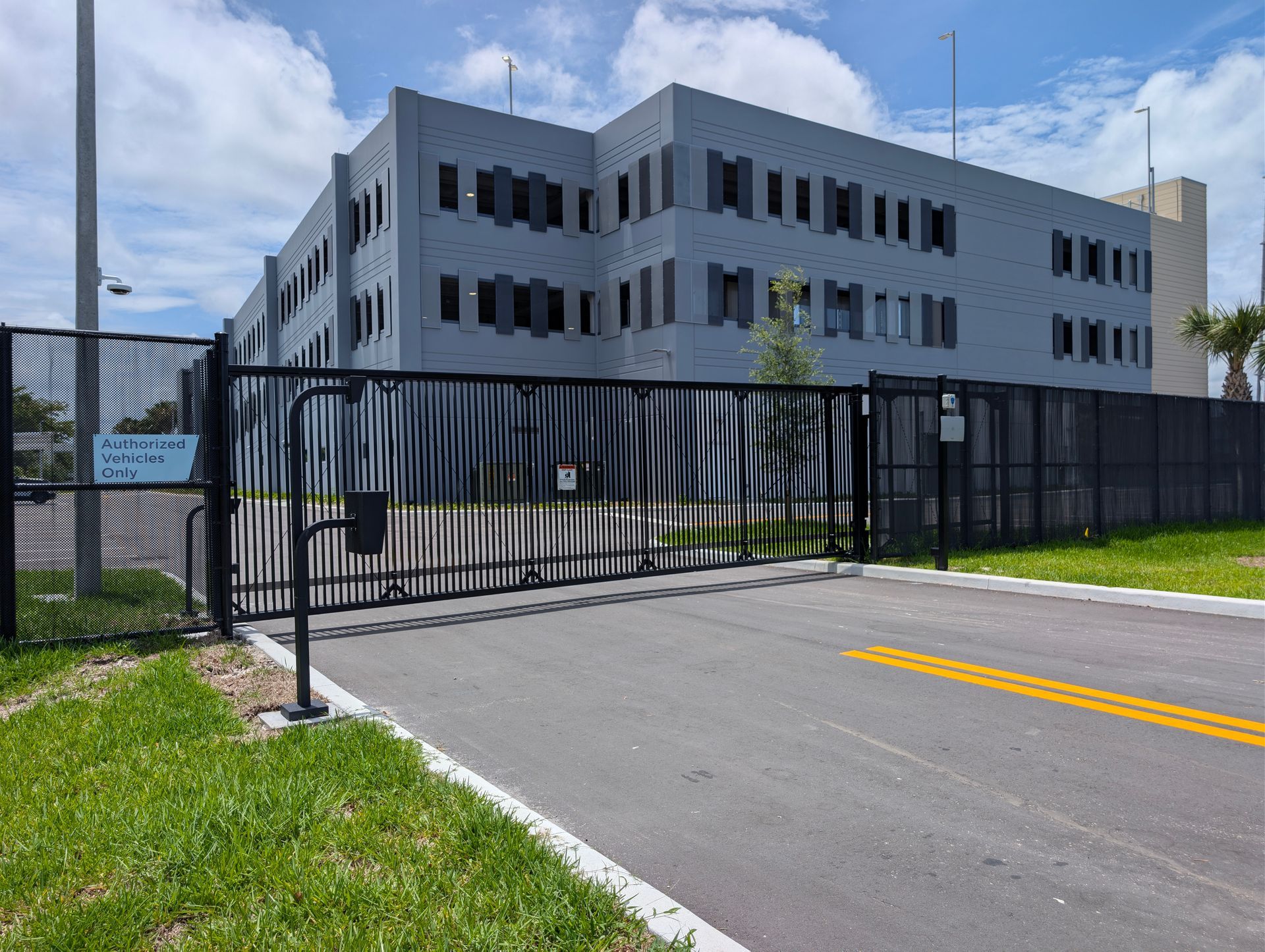 Modern gray office building behind a black metal gate and driveway under a blue sky