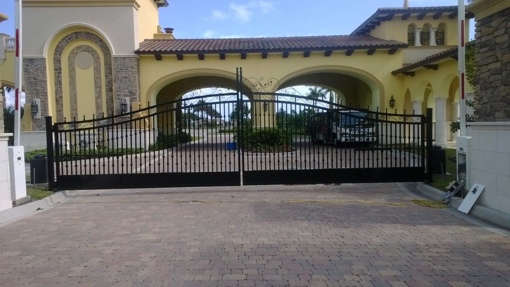 Black wrought-iron driveway gate in front of a yellow house with arched windows and tiled roof