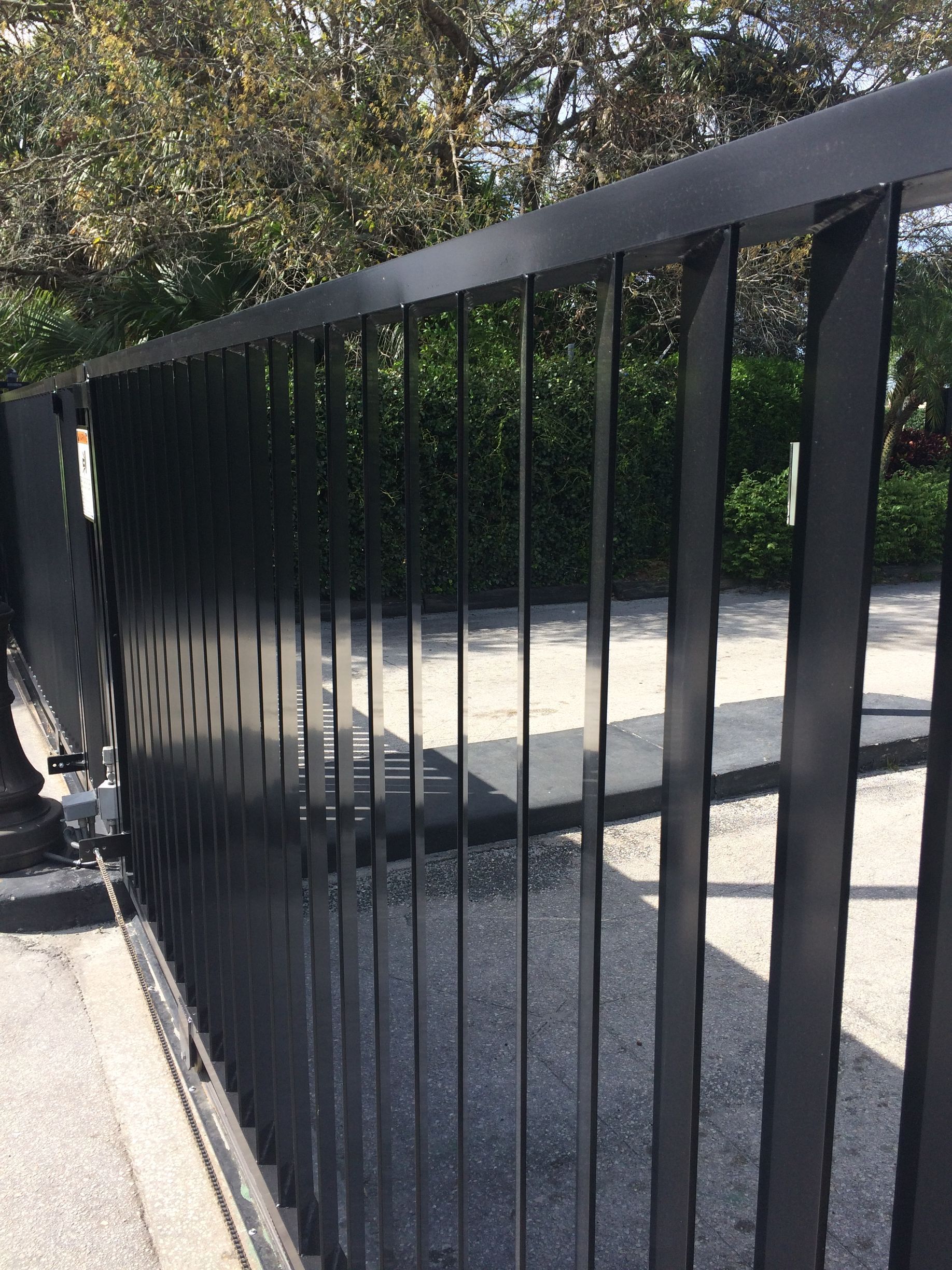 Black metal railing beside a sunlit concrete walkway with trees and shrubs in the background
