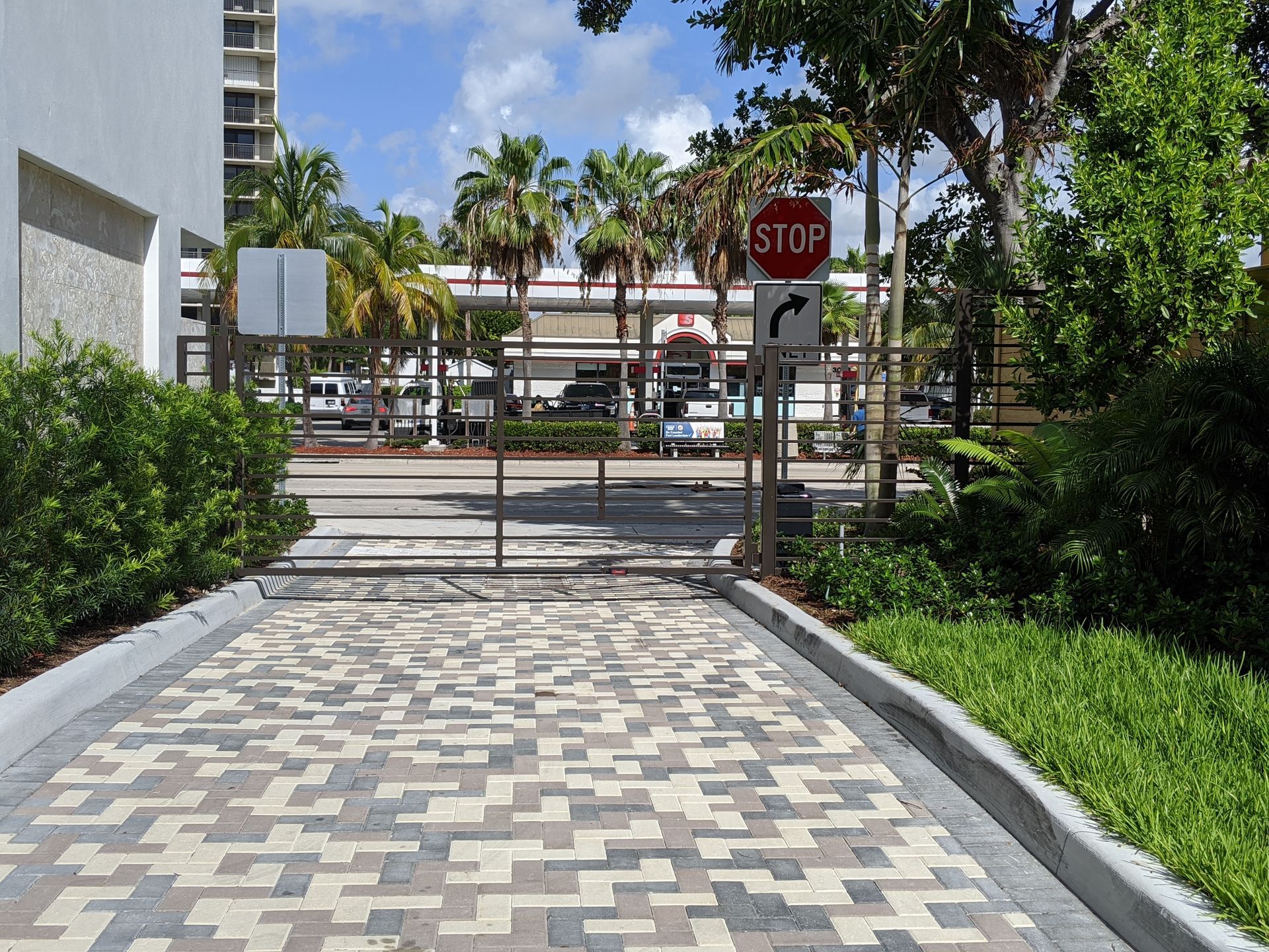 Paved walkway leading to a gated entrance with a stop sign, palm trees, and buildings in the background