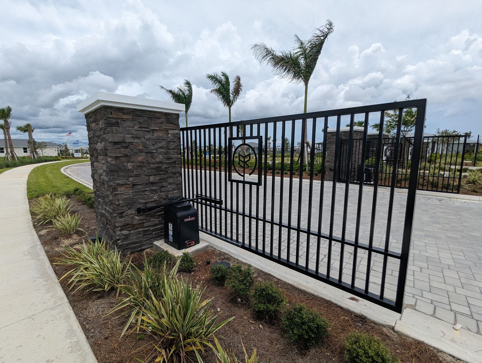 Black metal gate beside a stone pillar and landscaped walkway under cloudy skies