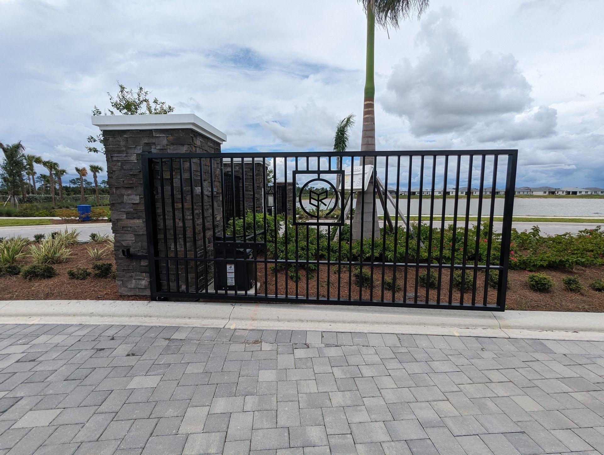 Black metal gate and stone pillar beside a landscaped waterfront walkway under cloudy skies