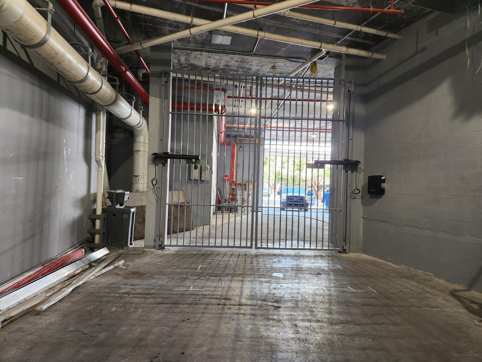 Industrial garage interior with concrete floor, exposed pipes, and a metal security gate opening to a bright exterior