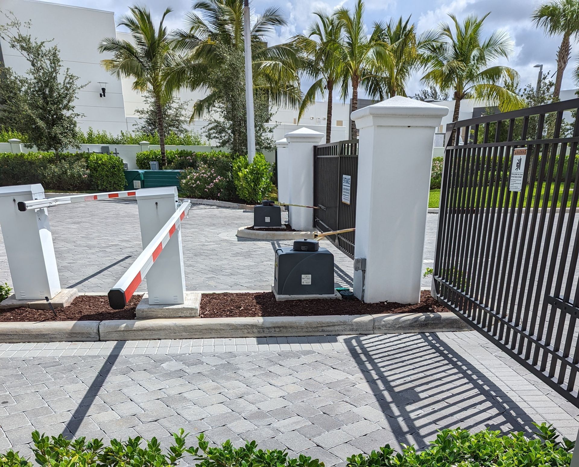 Gated entrance with barrier arm, gray pillars, and palm trees under bright sunlight