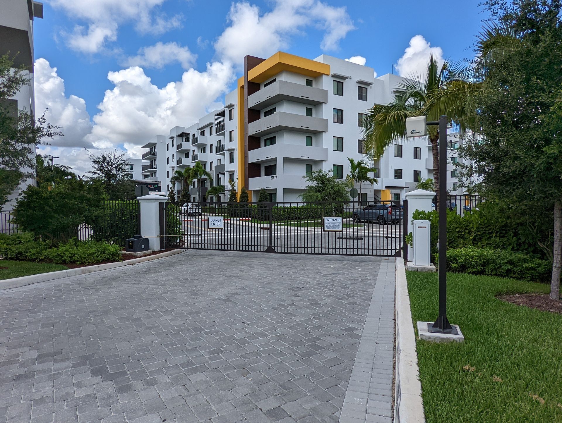 Gated entrance to a modern white apartment building with yellow accents, driveway, and green landscaping