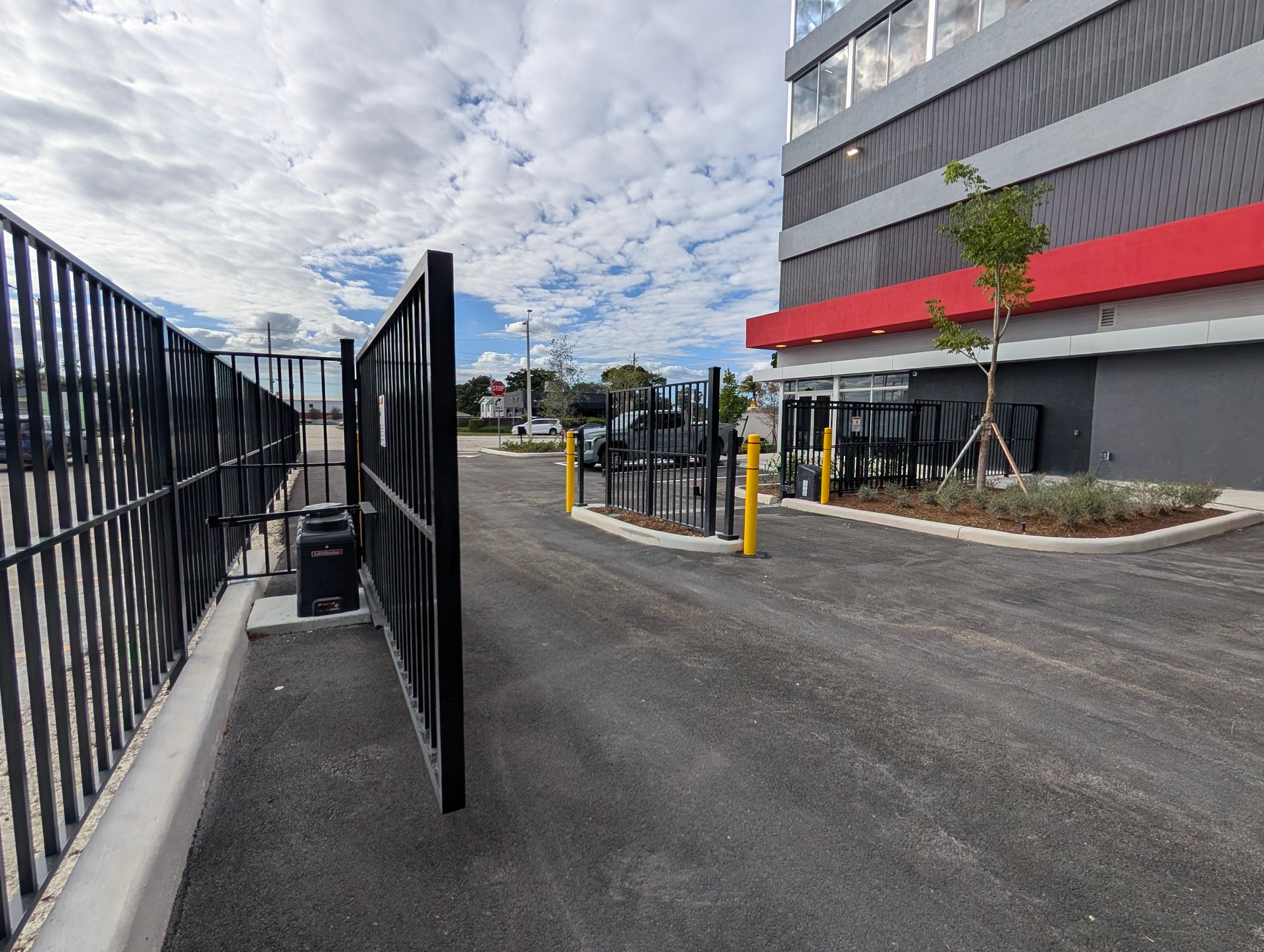 Gated driveway beside a modern building with yellow bollards and cloudy sky.