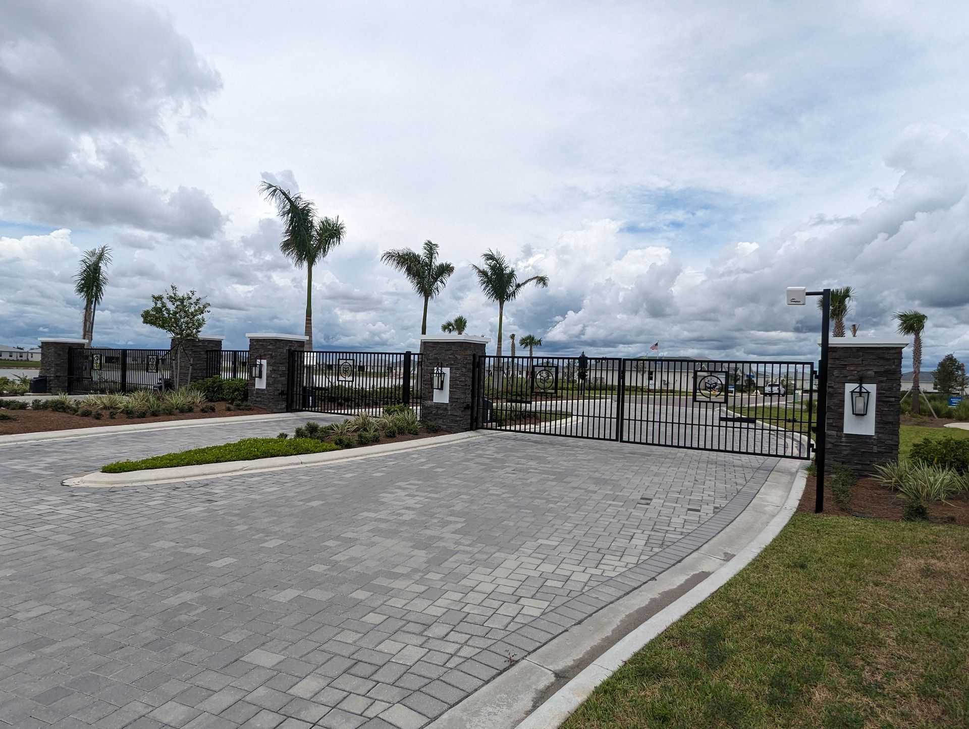 Gated entrance with stone driveway, palm trees, and cloudy sky at a modern residential or resort property