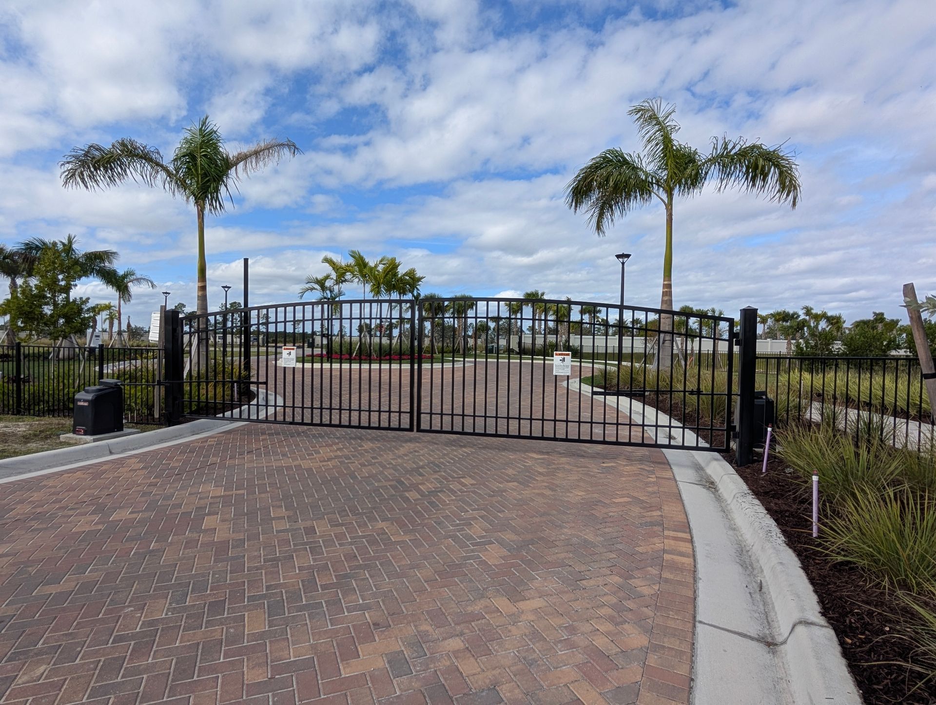 Gated brick driveway entrance with palm trees and a cloudy sky beyond
