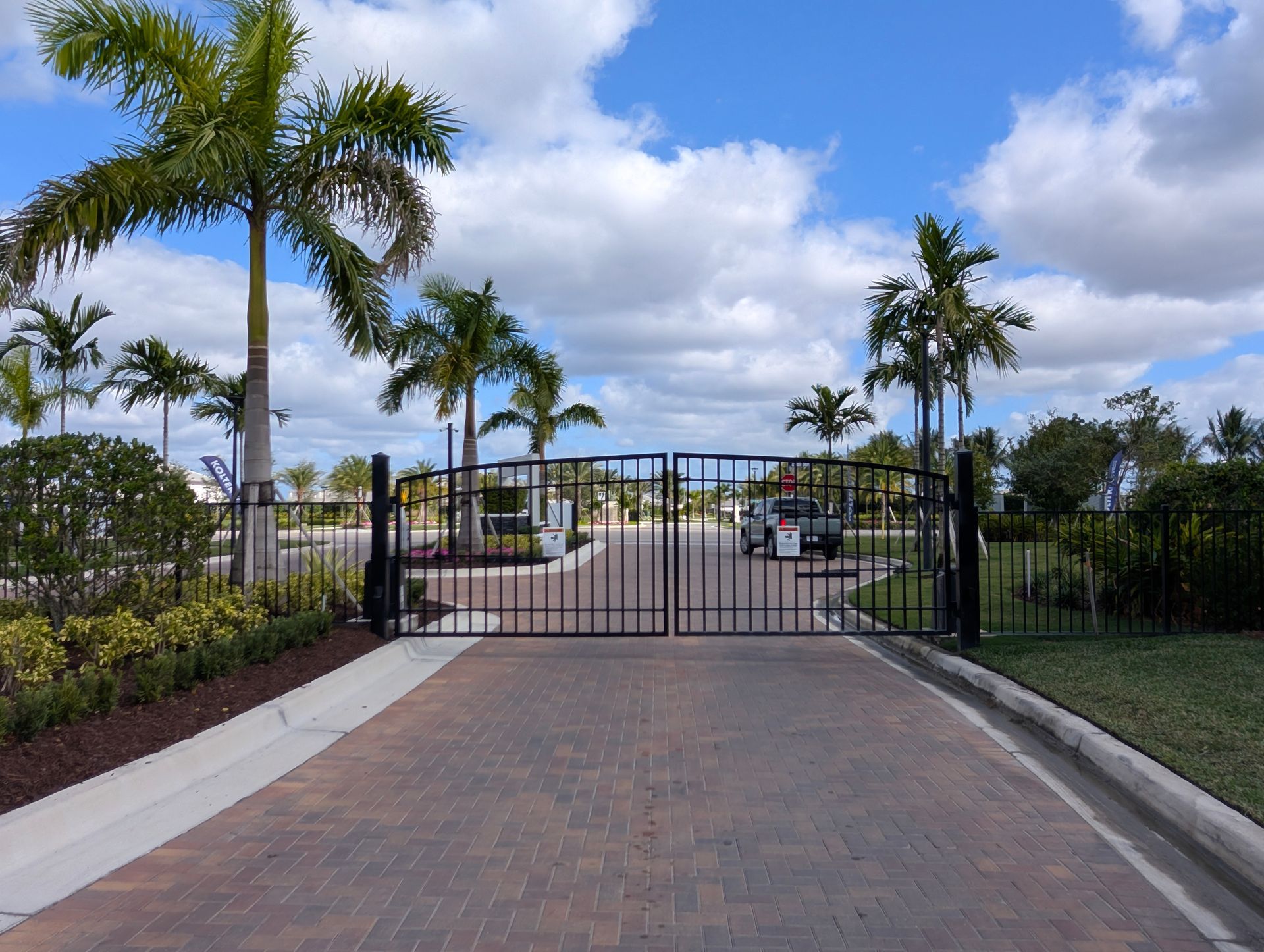 Gated entrance with brick driveway, palm trees, and a blue sky with clouds