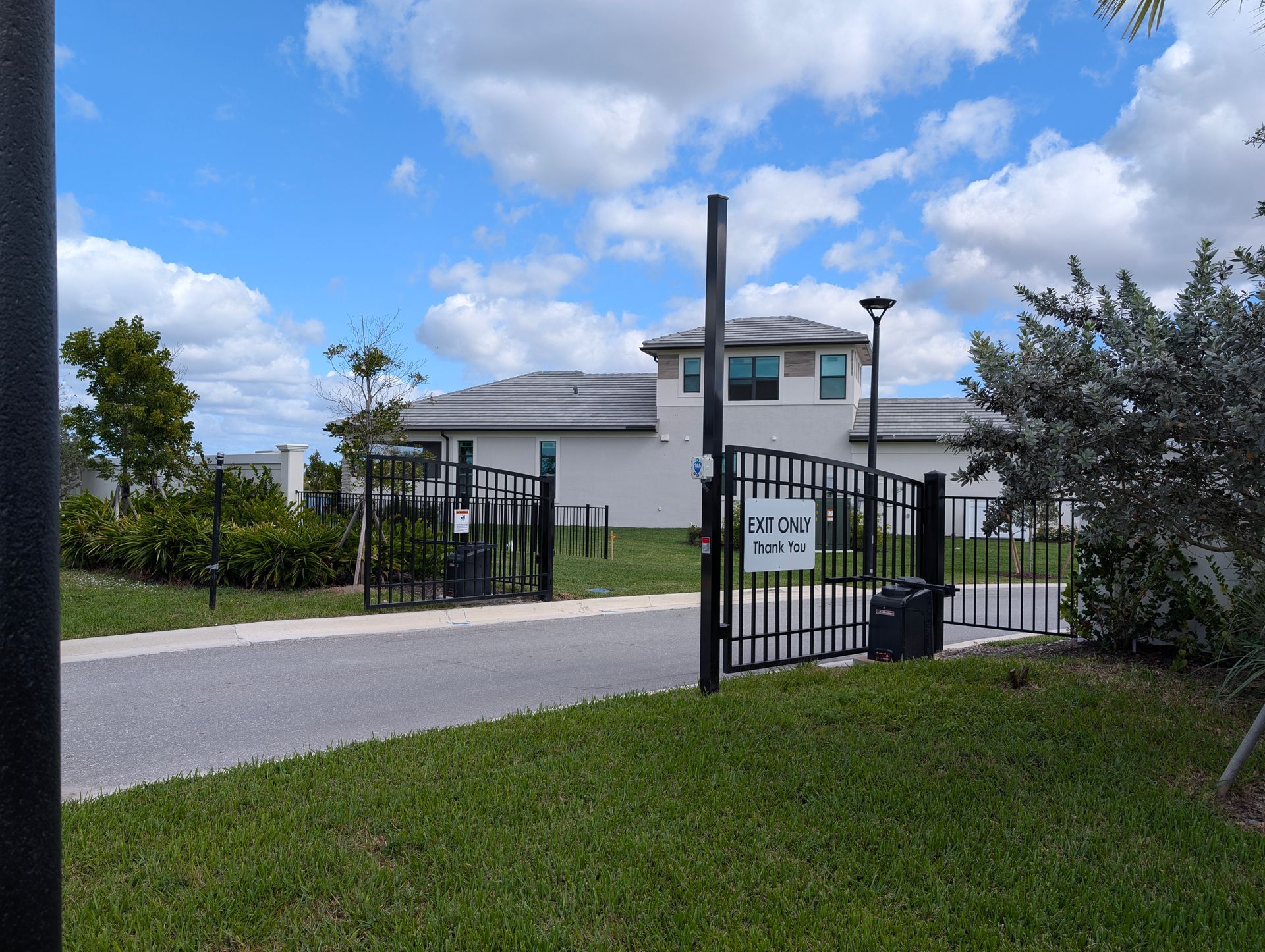 Gated entrance to a white building with a tower, black fence, driveway, and green lawn under a cloudy sky