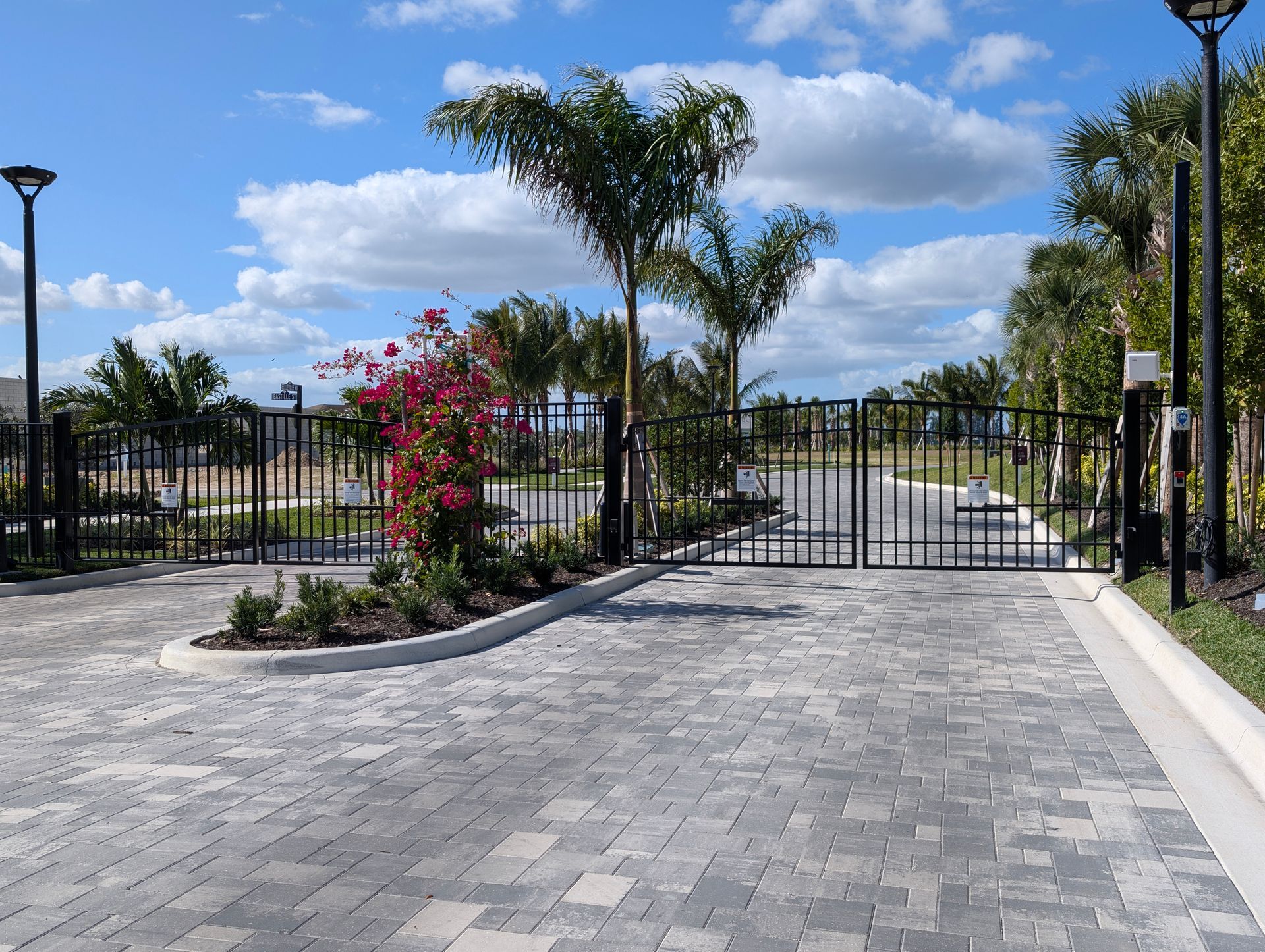 Paved driveway leading to a gated entrance with palm trees and flowering shrubs under a blue sky
