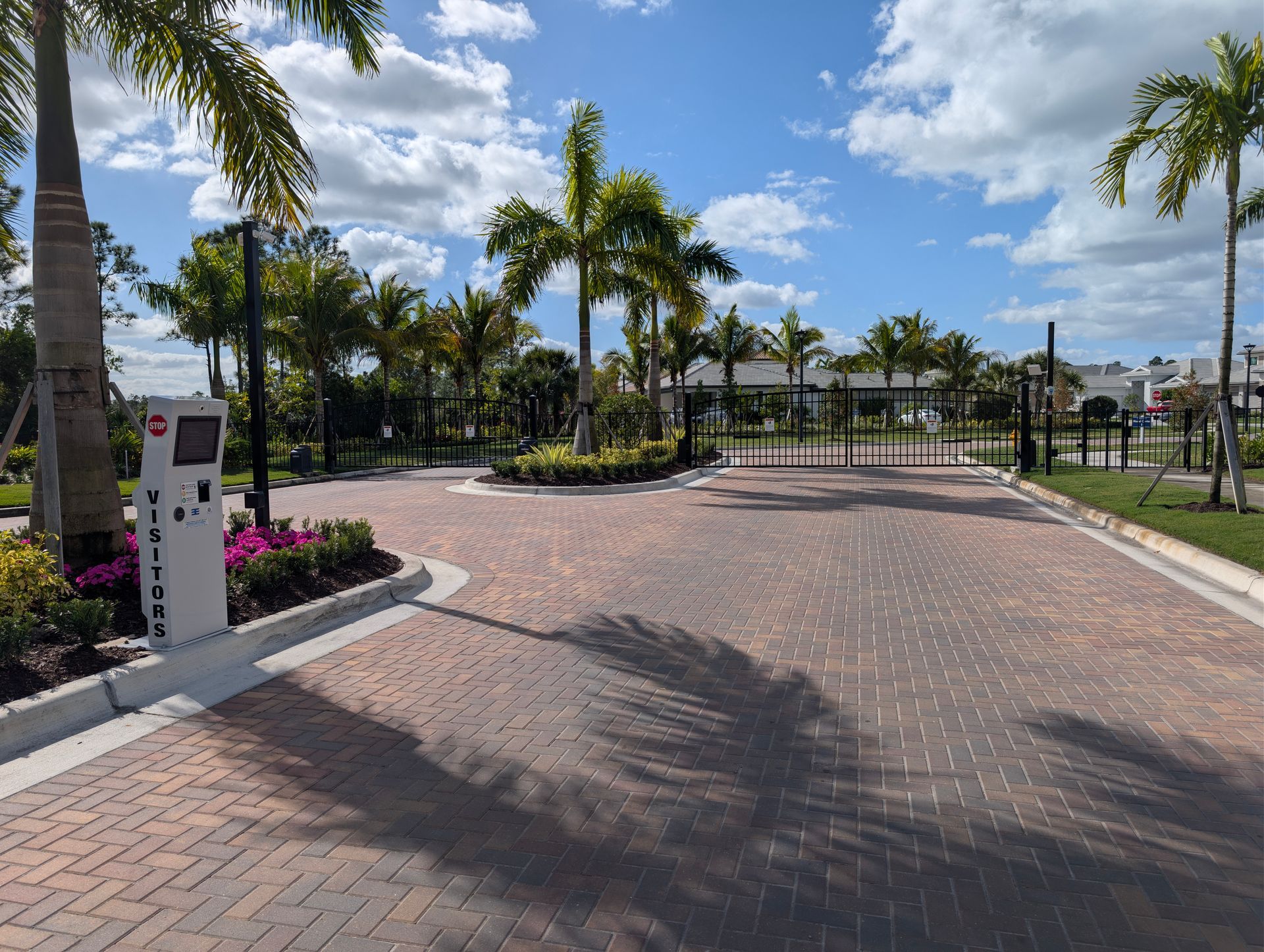 Brick-paved entrance with palm trees, flower beds, and a gated pathway under a bright blue sky