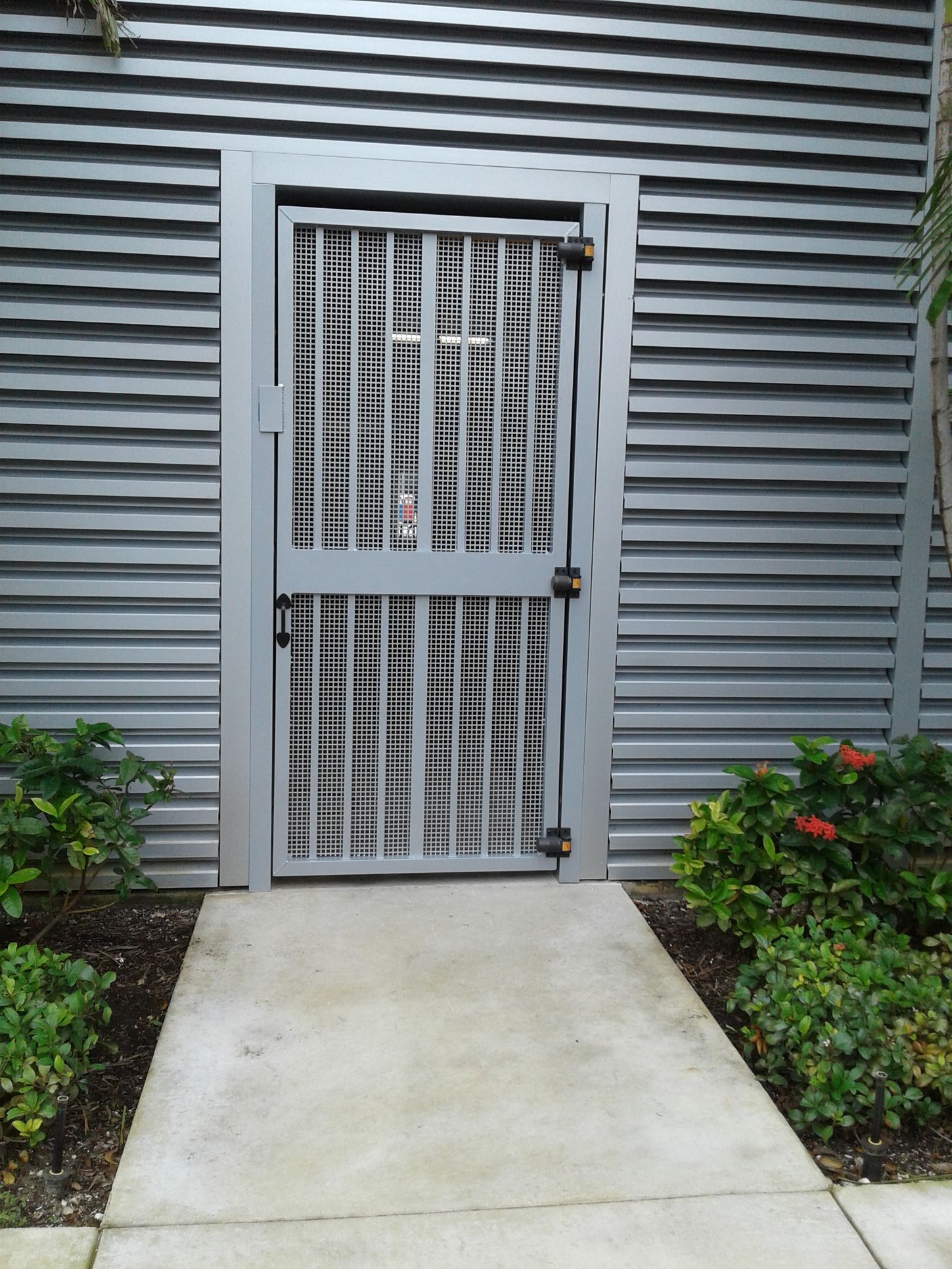 Gated doorway in a gray siding wall beside a concrete path and low shrubs