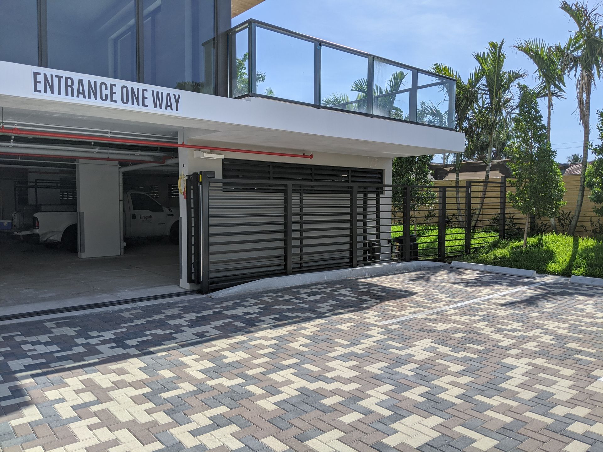 One-way entrance to a modern parking area with paved driveway, metal gate, and greenery beside a white building