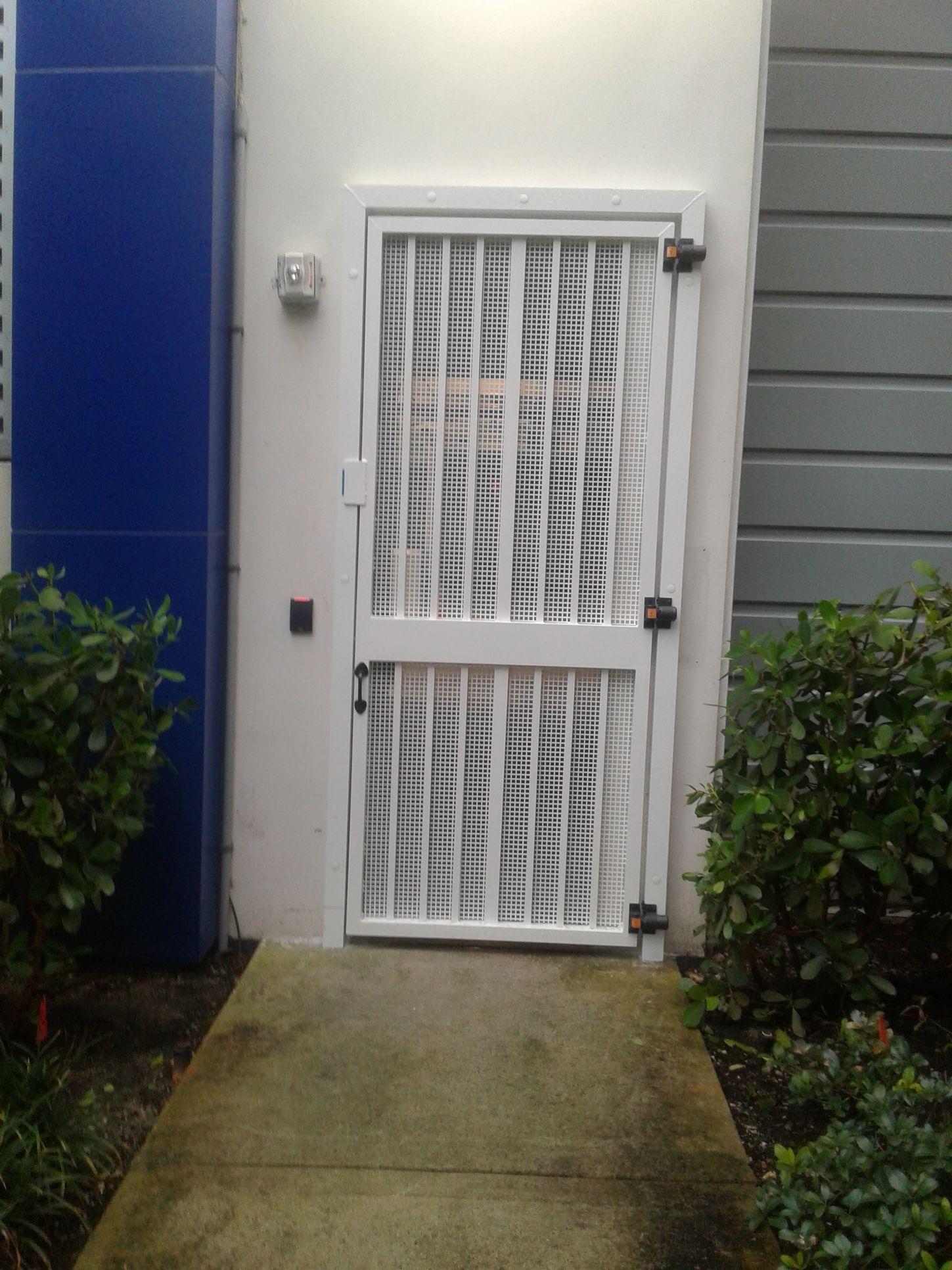 White security door with metal bars at the end of a narrow concrete walkway beside blue and gray siding.