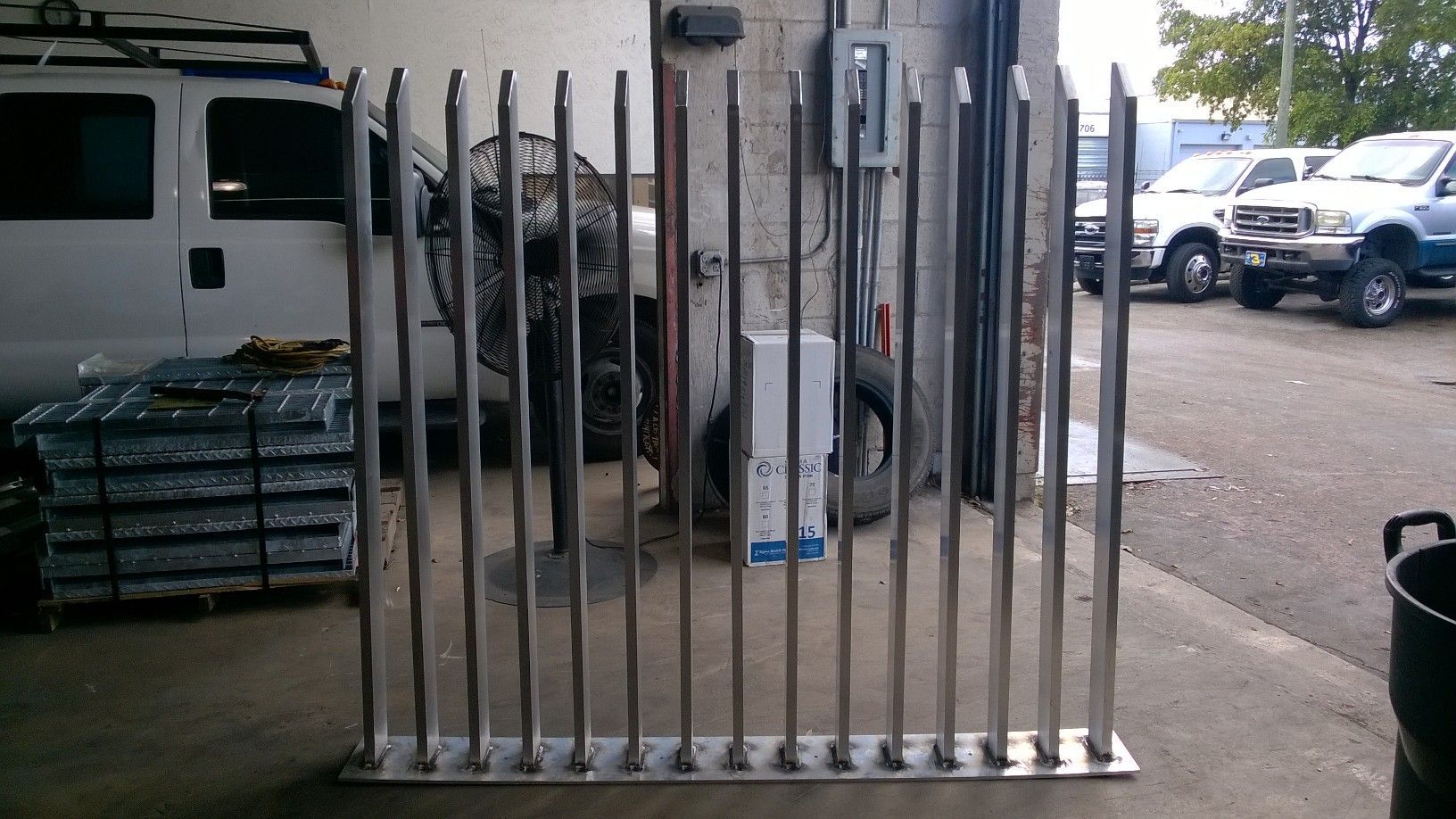 Metal barricade with vertical bars standing on a concrete lot beside a white van and parked trucks.