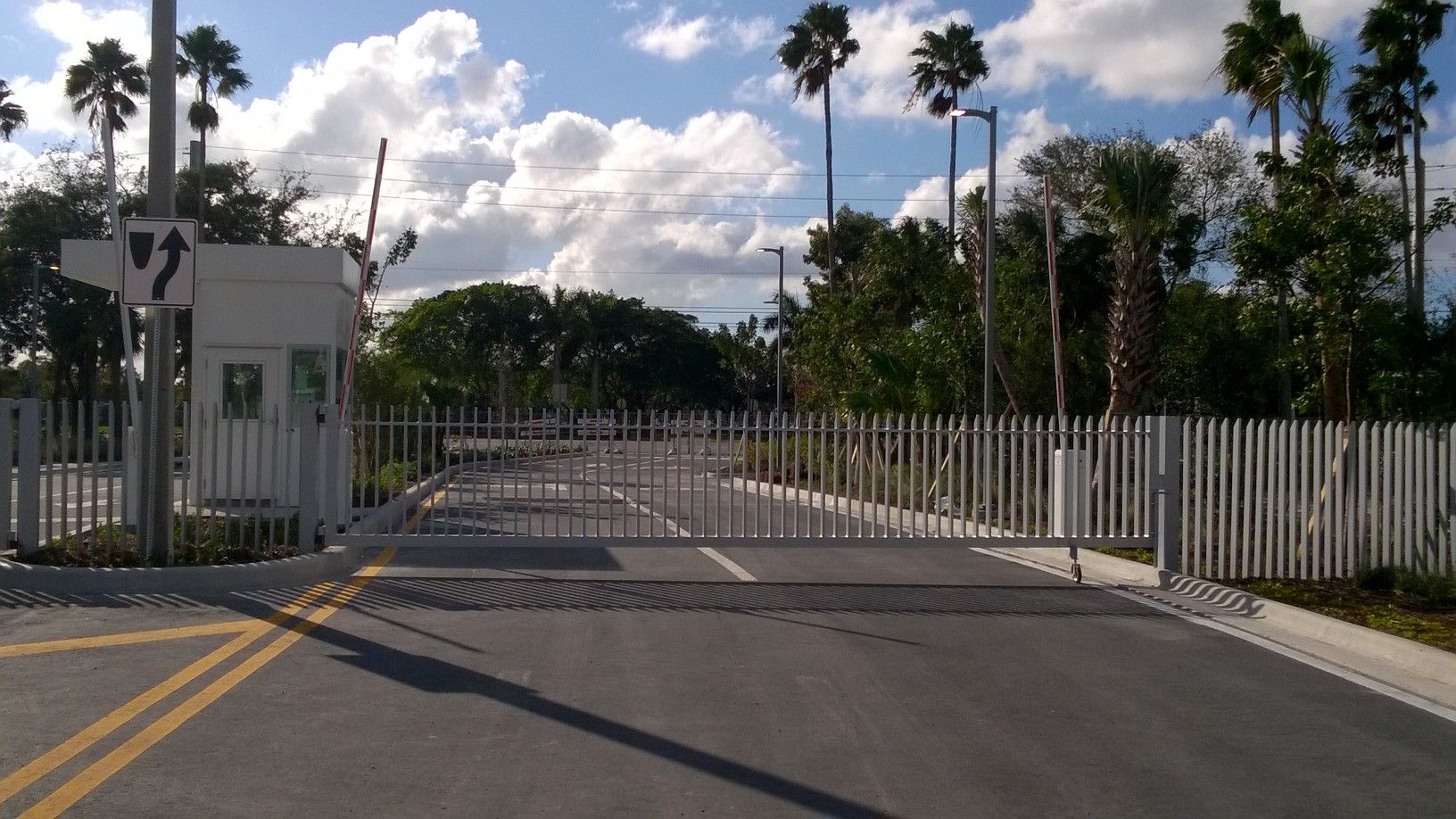 Closed gated entrance with white fence and palm trees under a cloudy sky