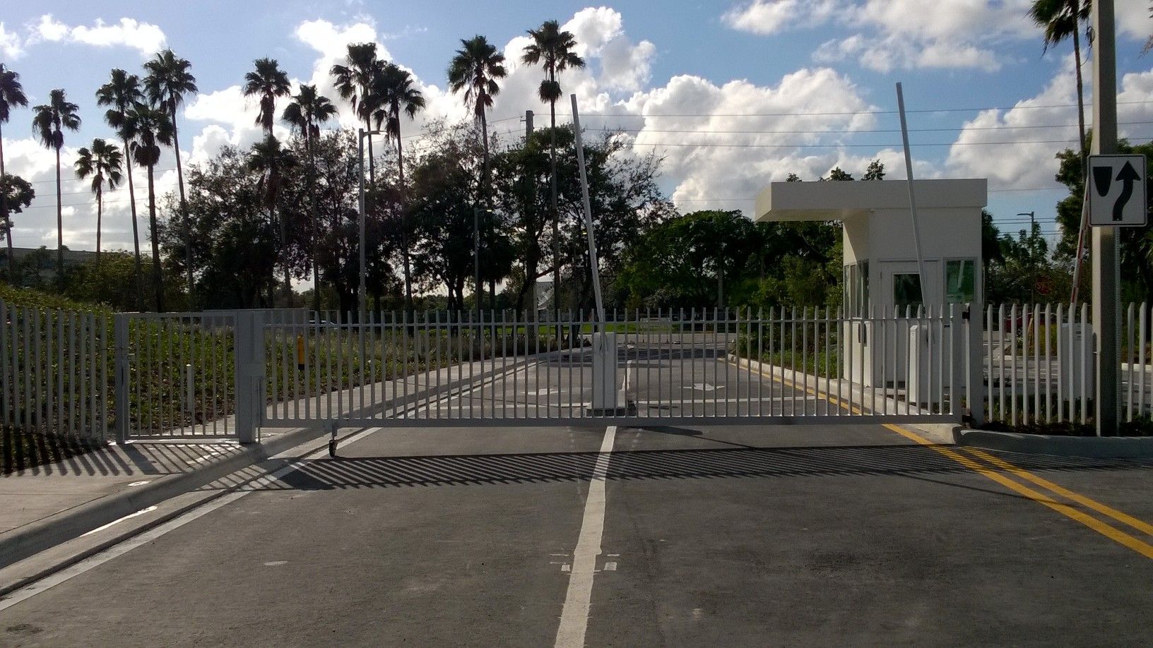 Gated road entrance with white fence, guard booth, and palm trees under a cloudy sky