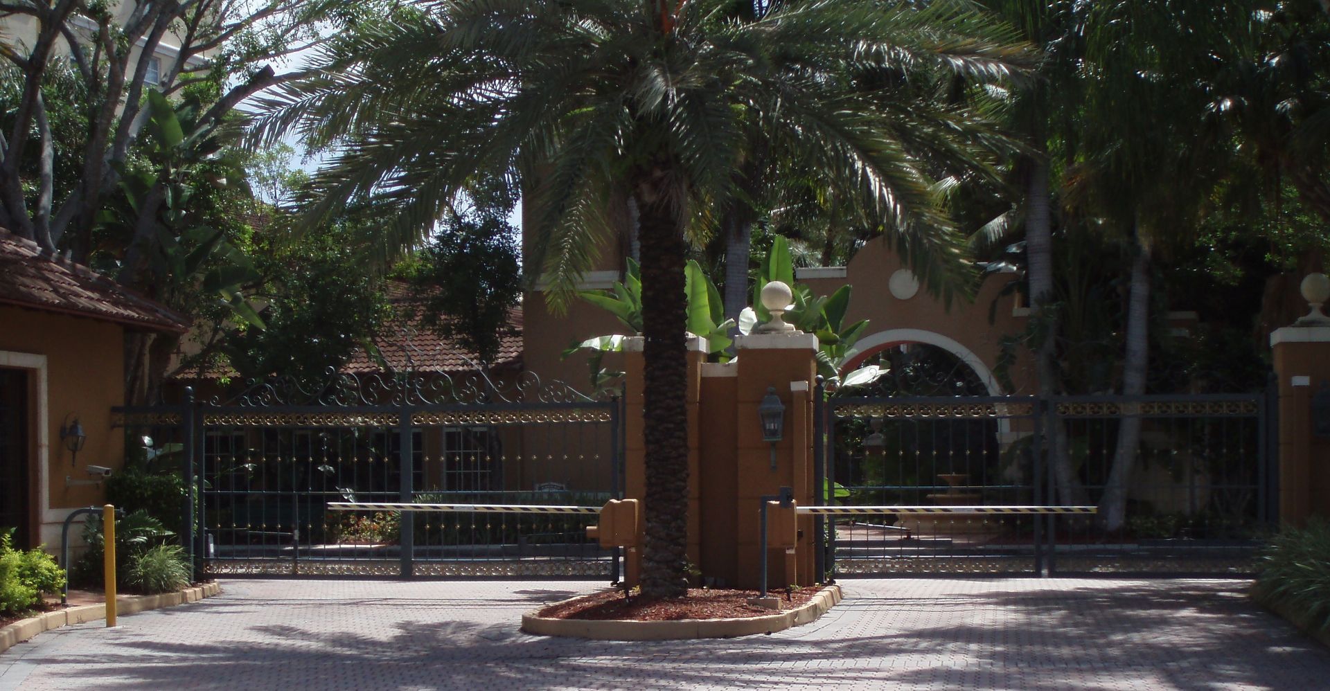 Gated entrance to a tropical home with palm trees, a brick pillar, and a metal gate in sunlight
