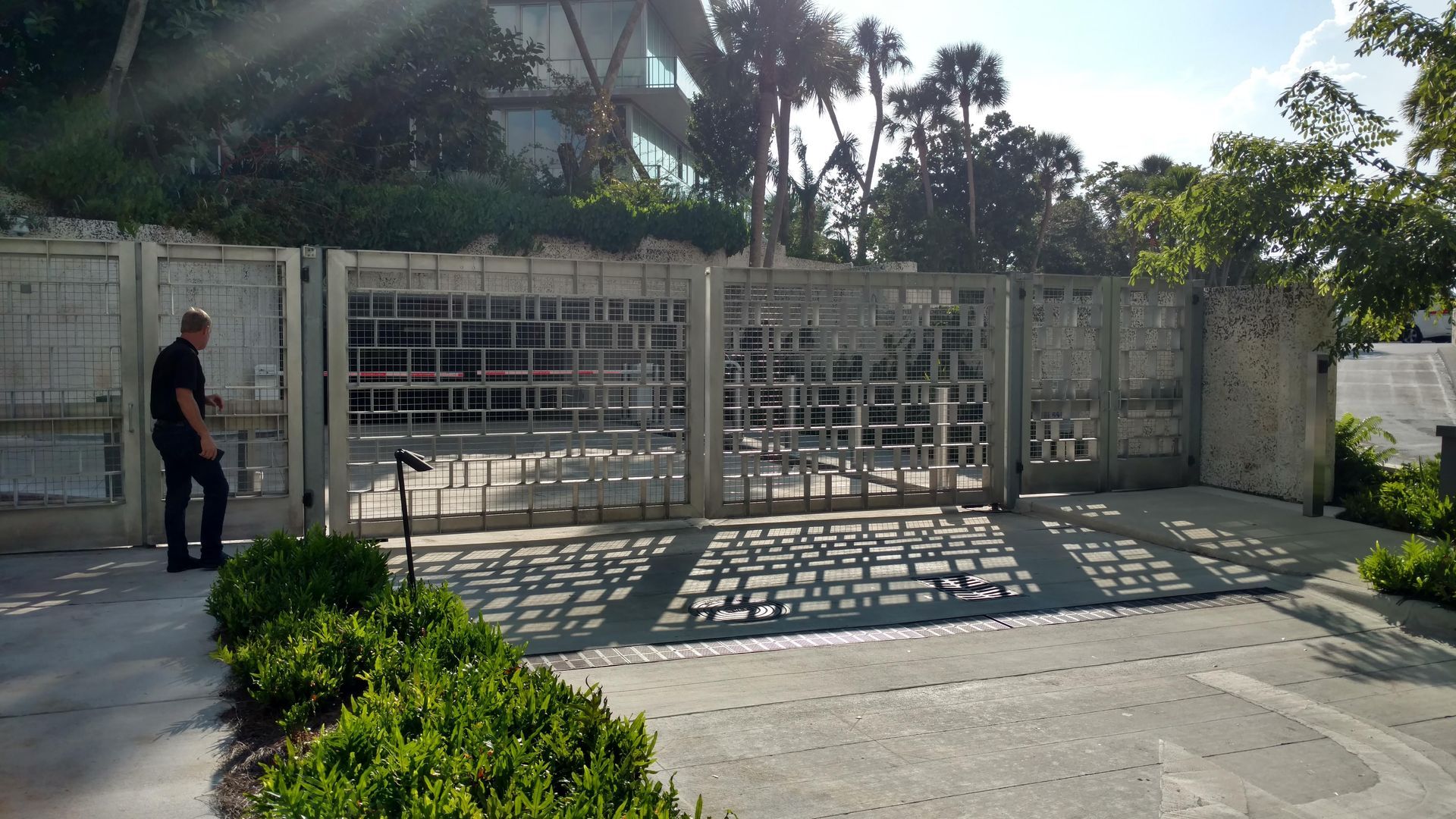 Person standing beside a closed metal gate in front of a sunlit residential driveway