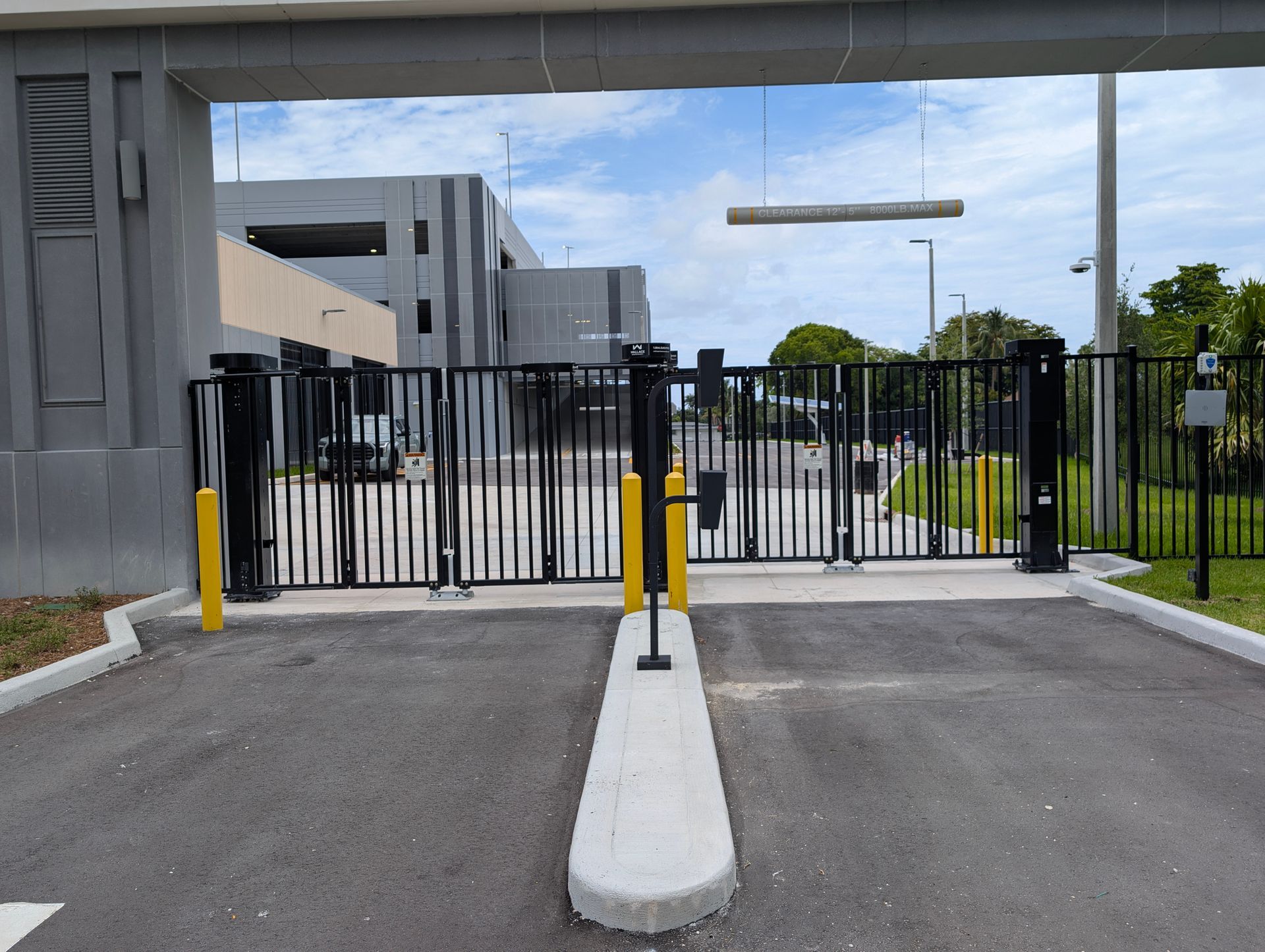 Closed black metal gate blocking a driveway beside a gray building, with yellow bollards and a concrete divider.
