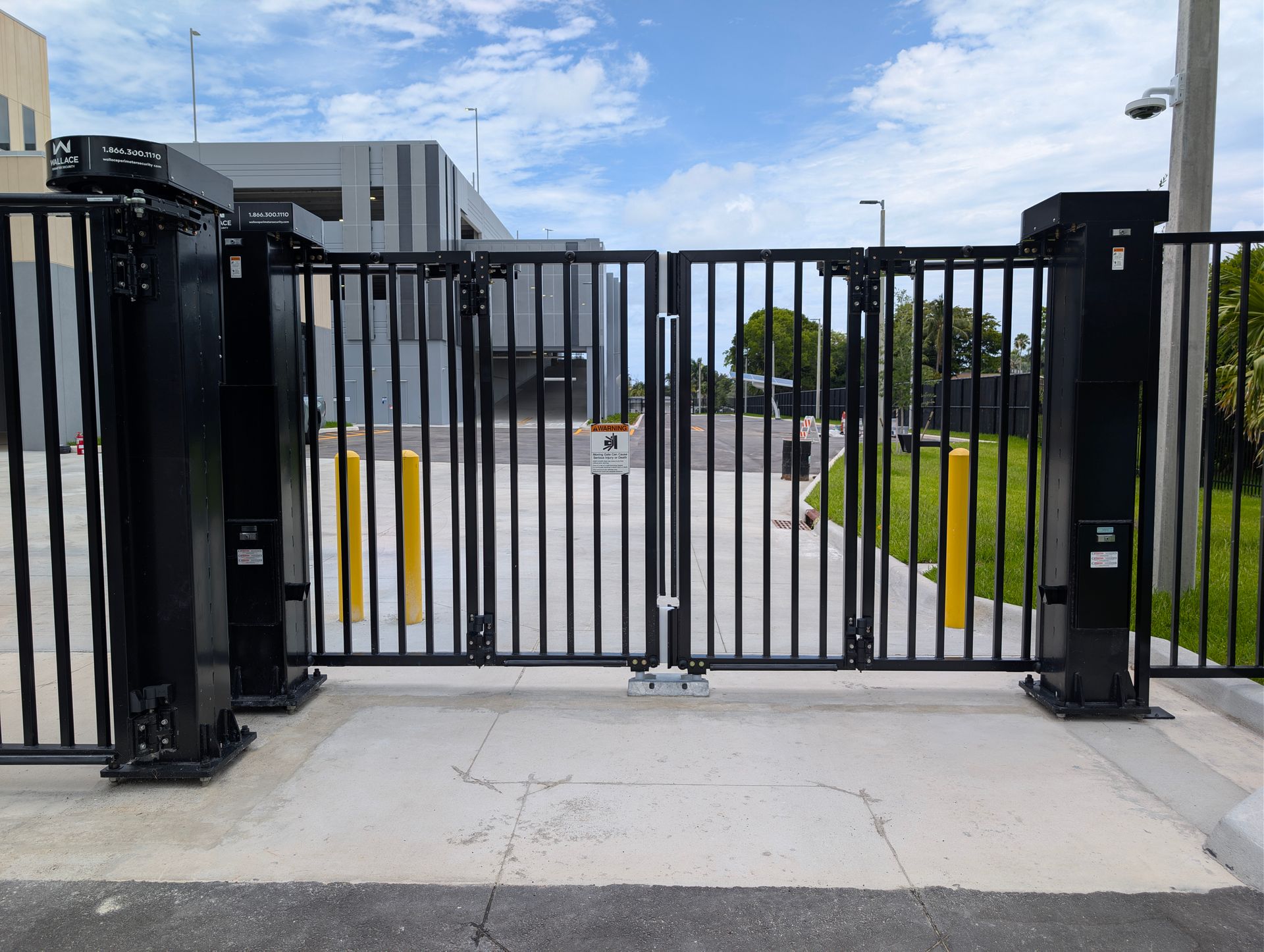 Black metal security gate across a paved entrance with buildings and grass beyond.