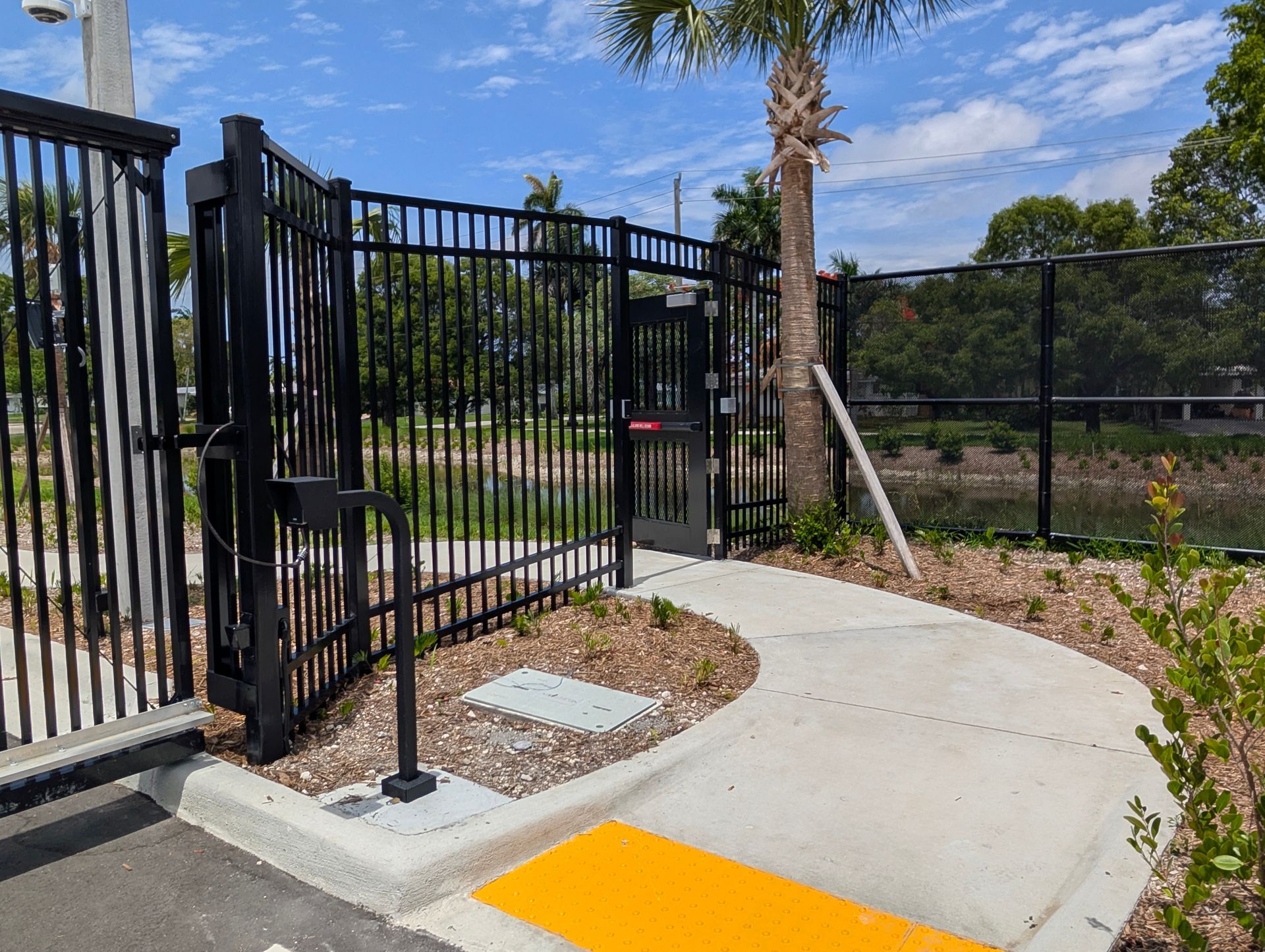 Accessible gated walkway with curved concrete path, black fence, and yellow tactile curb cut under blue sky