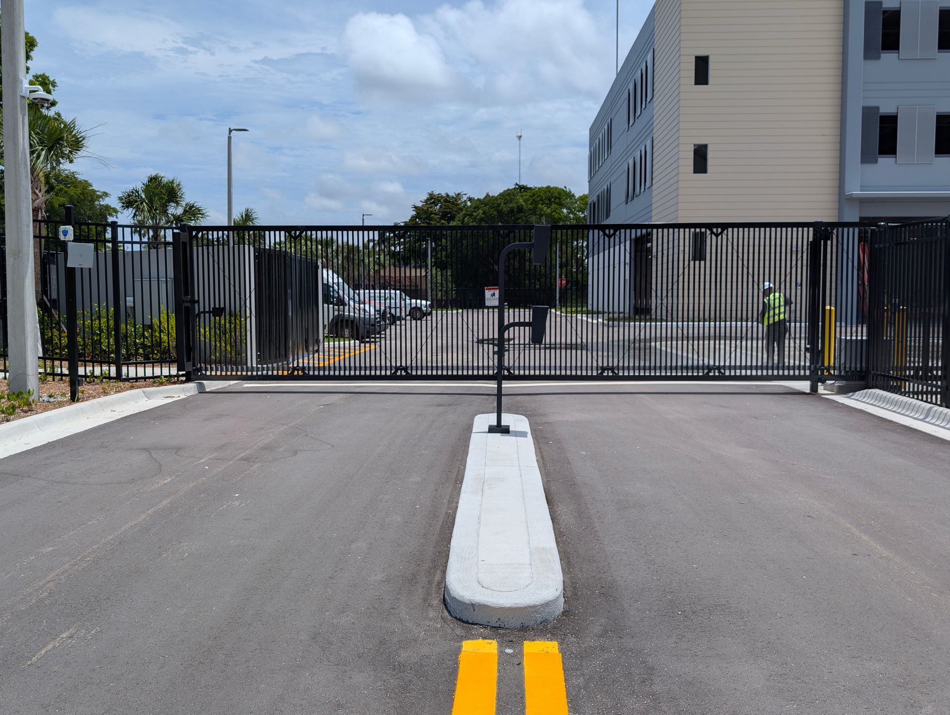 Closed black gate blocking a driveway entrance beside a white building under a partly cloudy sky